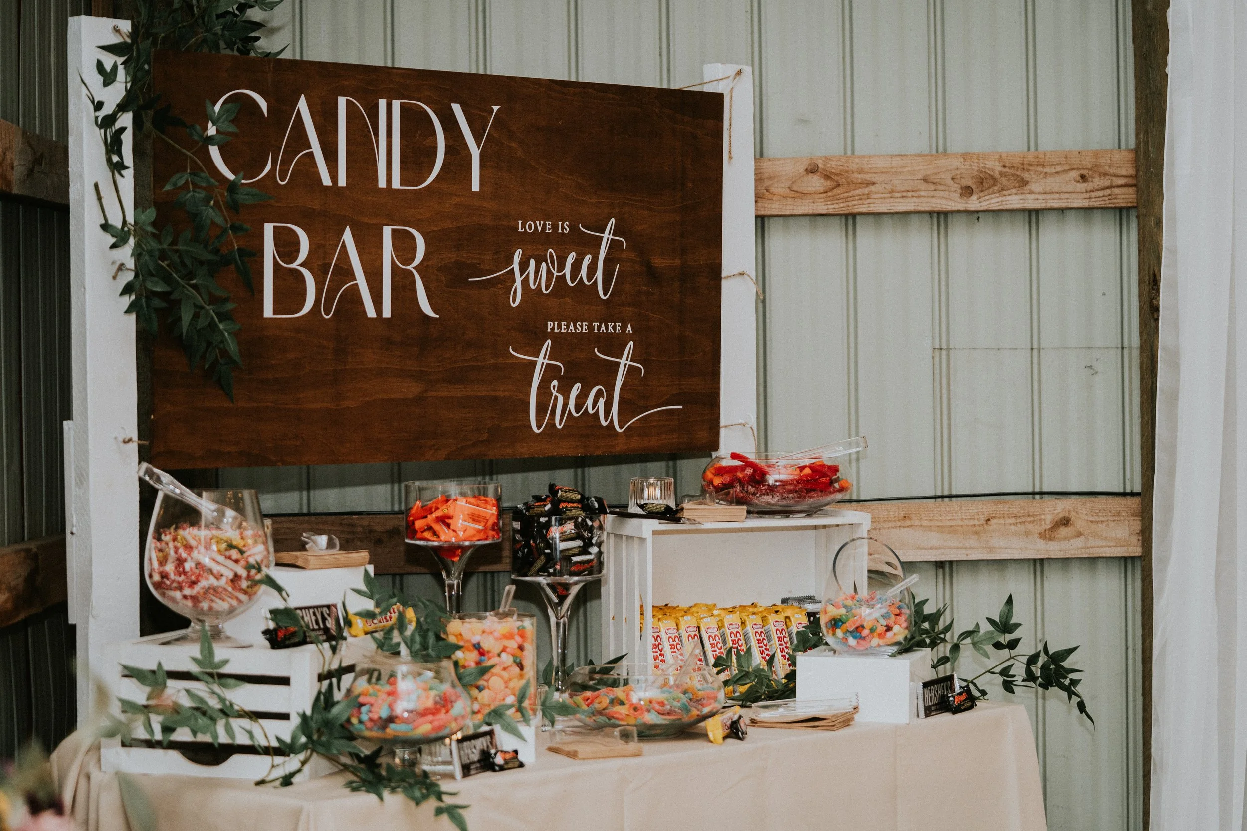 A candy bar station with a wooden sign that says 'Candy Bar' and 'Love is sweet, please take a treat'. The table has various jars and bowls filled with colorful candies, surrounded by green leaves for decoration.