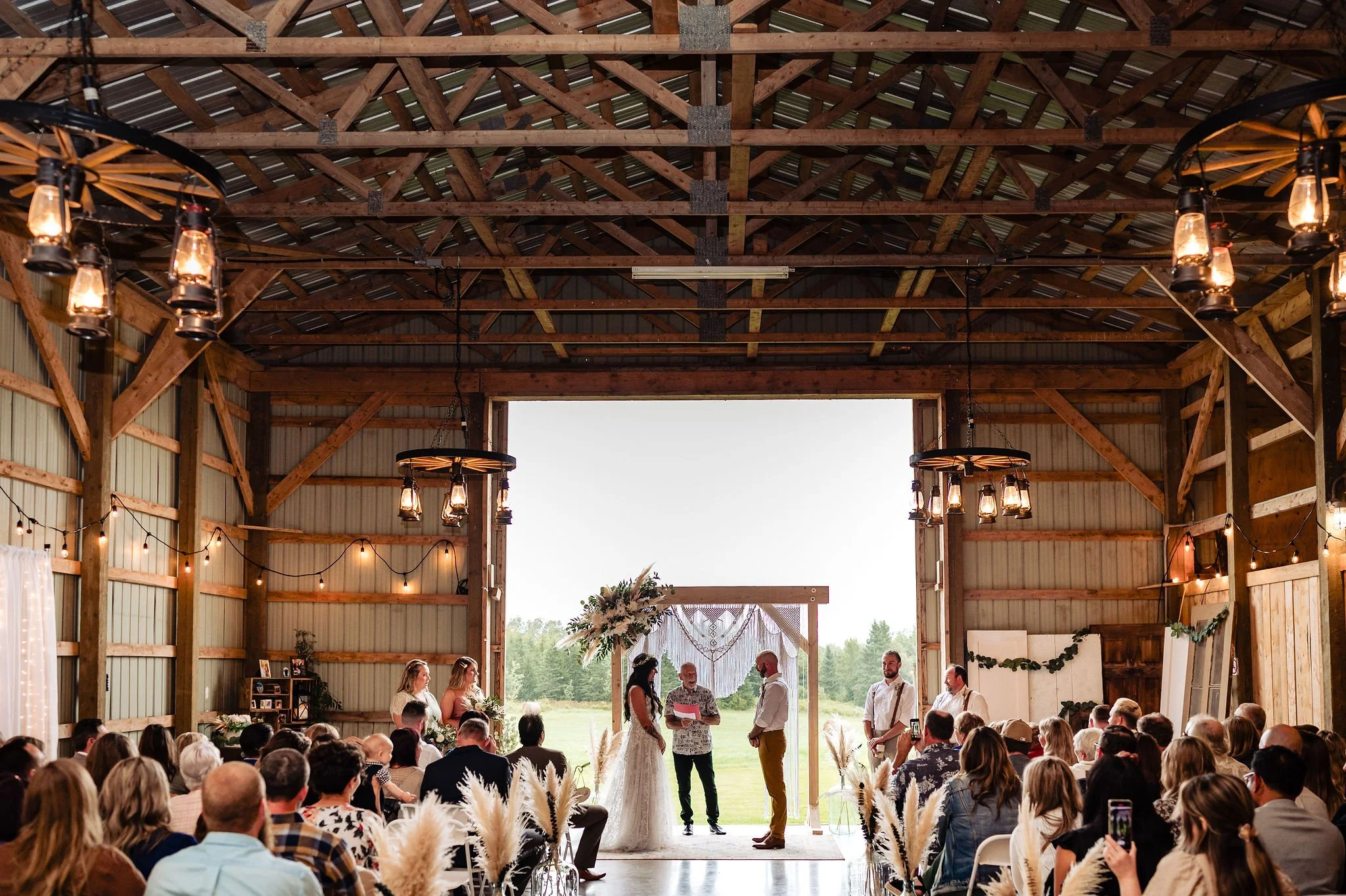 A wedding ceremony taking place inside a rustic barn with wood beams and hanging lanterns, with a bride and groom standing at an altar in front of officiants, surrounded by seated guests.