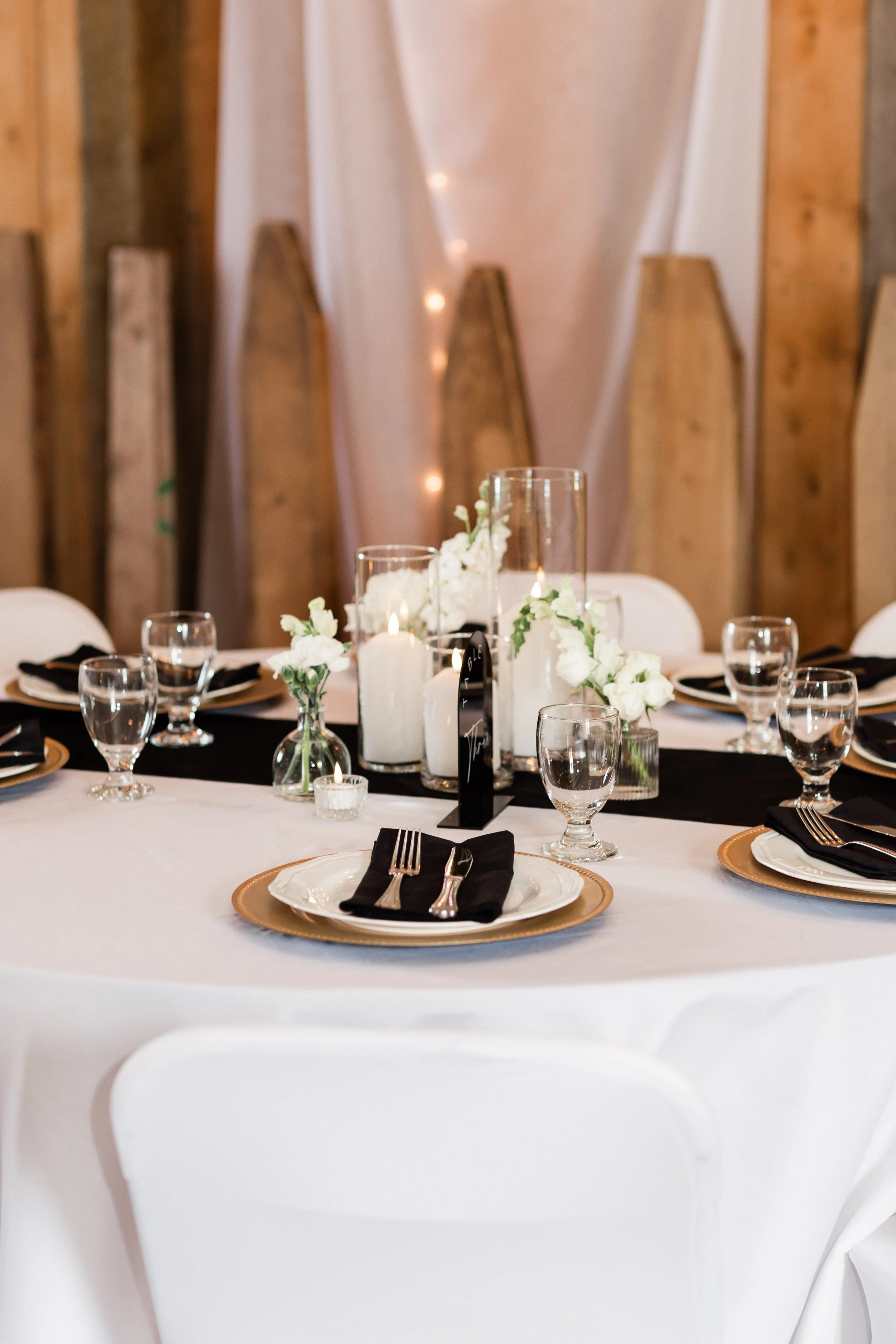 Elegant round dining table set for a formal event with white tablecloth, black napkins, gold-rimmed plates, glassware, and floral centerpieces with candles, in a rustic decorated venue.