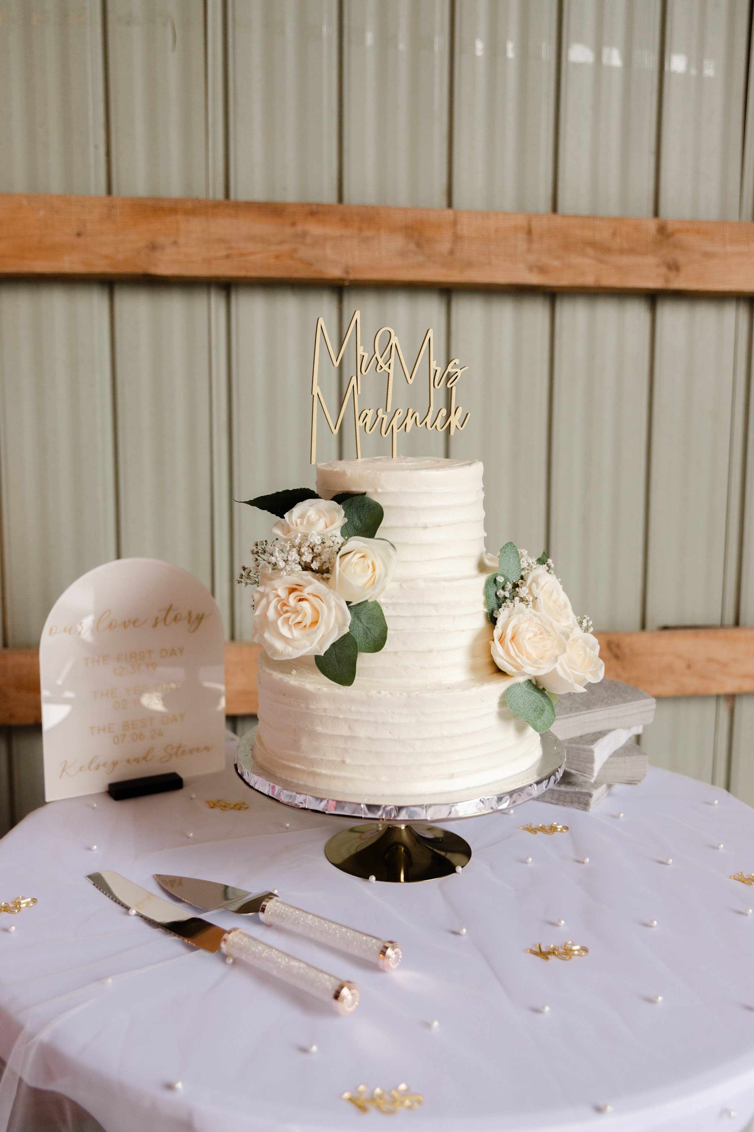 A wedding cake decorated with white roses and green leaves, topped with a gold cake topper that says 'Mr & Mrs Marenick.' The cake is on a round silver stand on a decorated table with cake serving utensils and small gold accents, set against a rustic