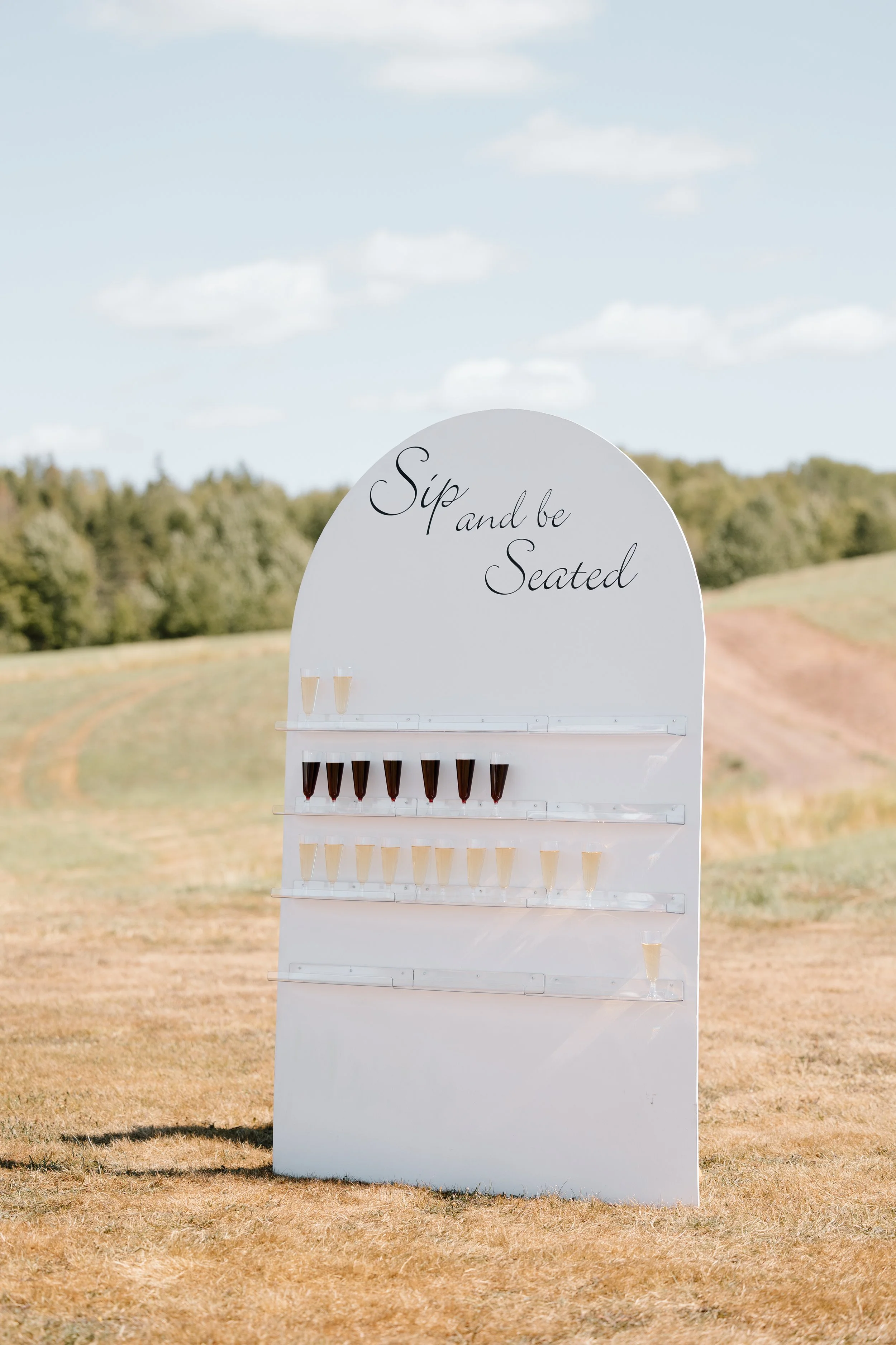 A white signboard with the words 'Sip and be Seated' written in cursive. The sign displays various cups of beverages, including red and light-colored drinks, arranged on horizontal transparent shelves outdoors on a grassy field with trees and a blue 