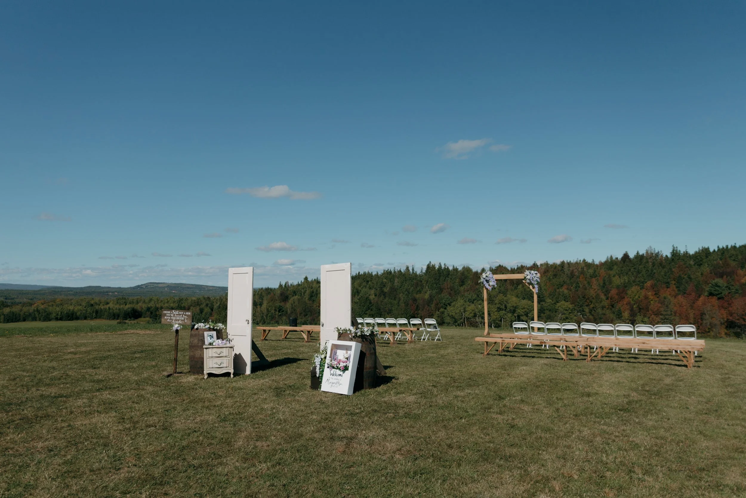 Outdoor wedding ceremony setup with white chairs, wooden arch decorated with flowers, and signs on a grassy field with distant hills and trees under a blue sky.