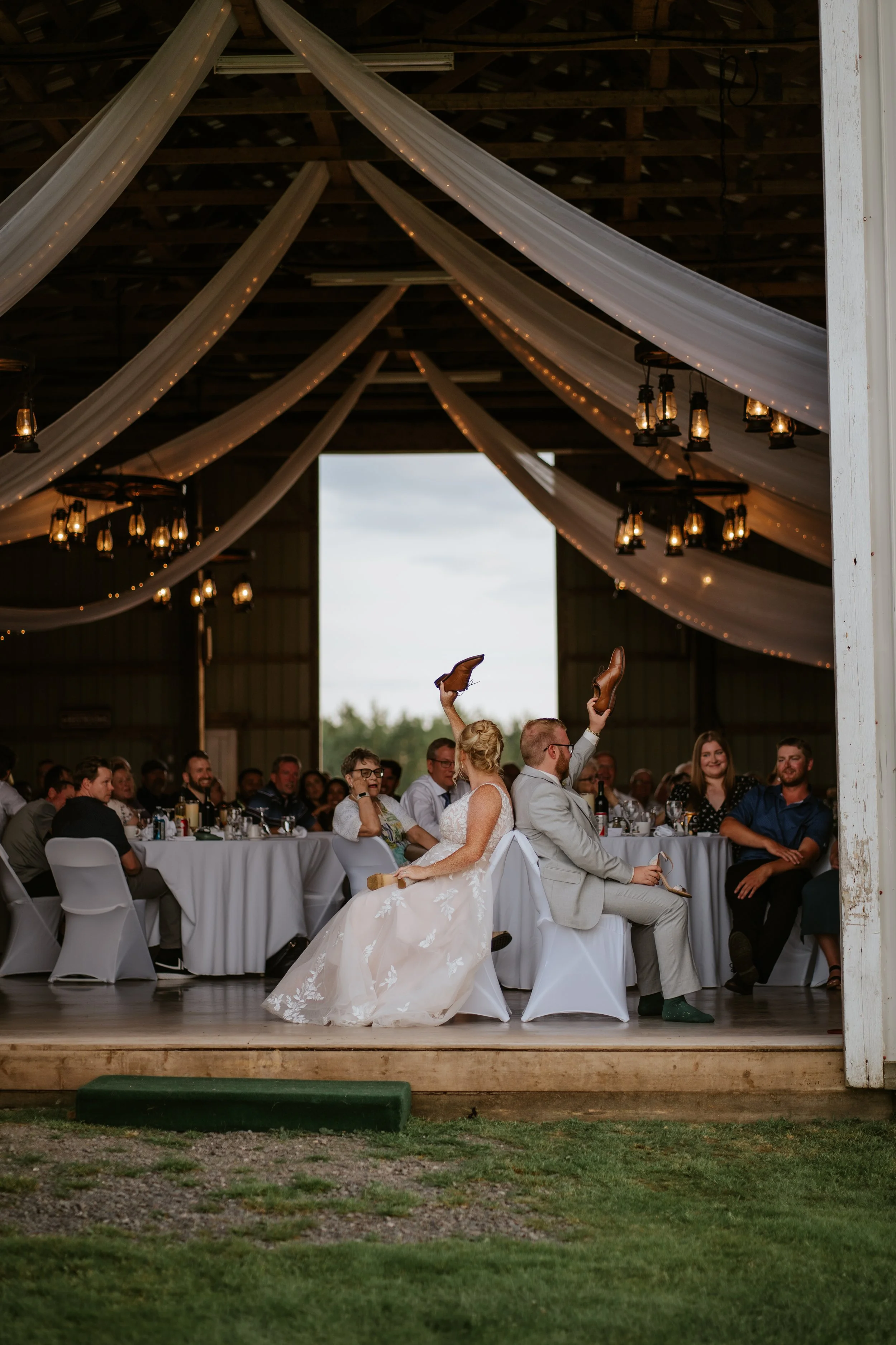 A wedding reception inside a barn. The bride and groom are sitting back-to-back in the center, raising their shoes as part of a wedding game, with bridesmaids and groomsmen behind them and guests seated at decorated tables. White drapes and string li