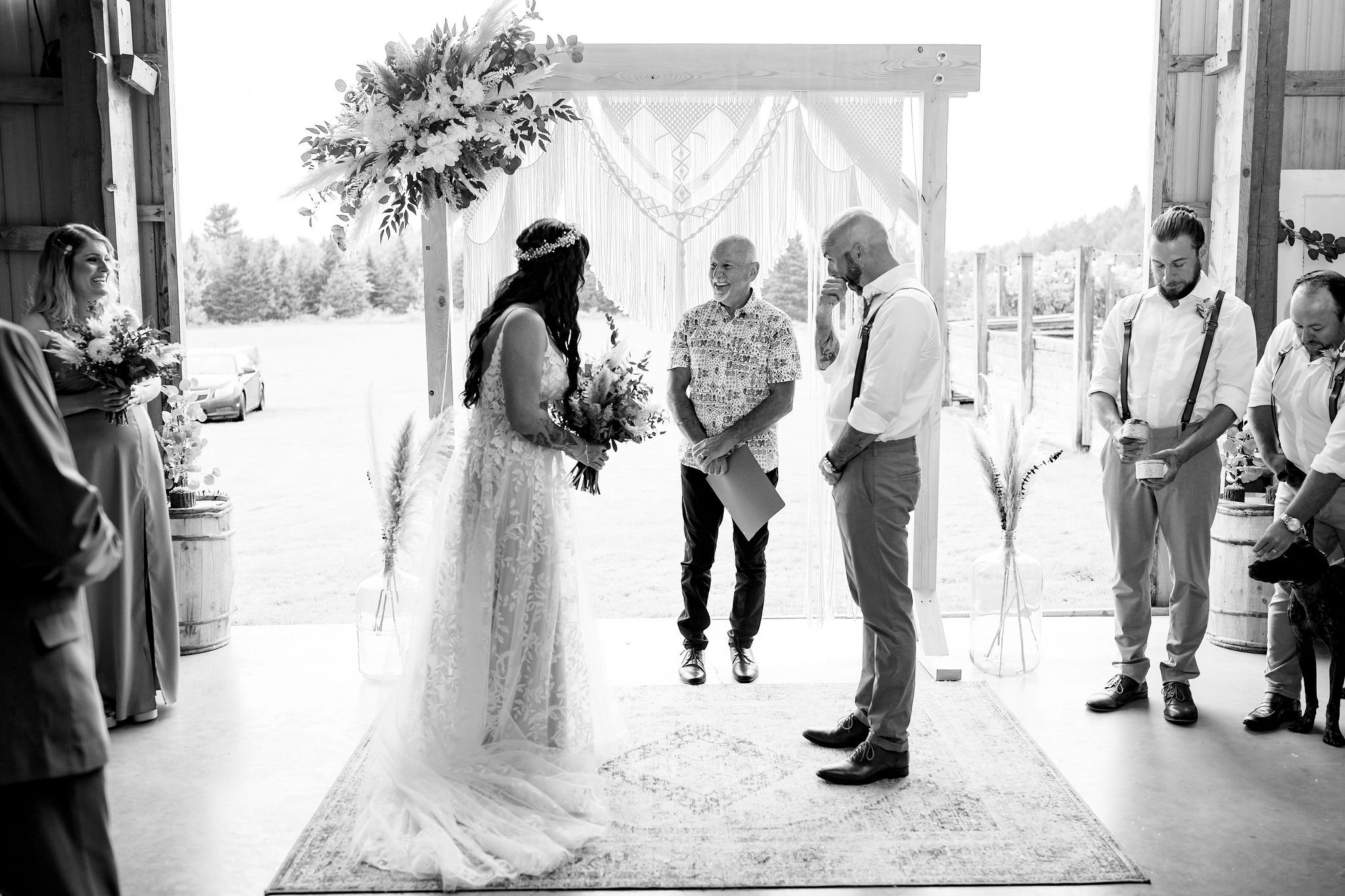 A bride and groom exchanging vows indoors at a rustic wedding venue, with officiant and wedding party members present.