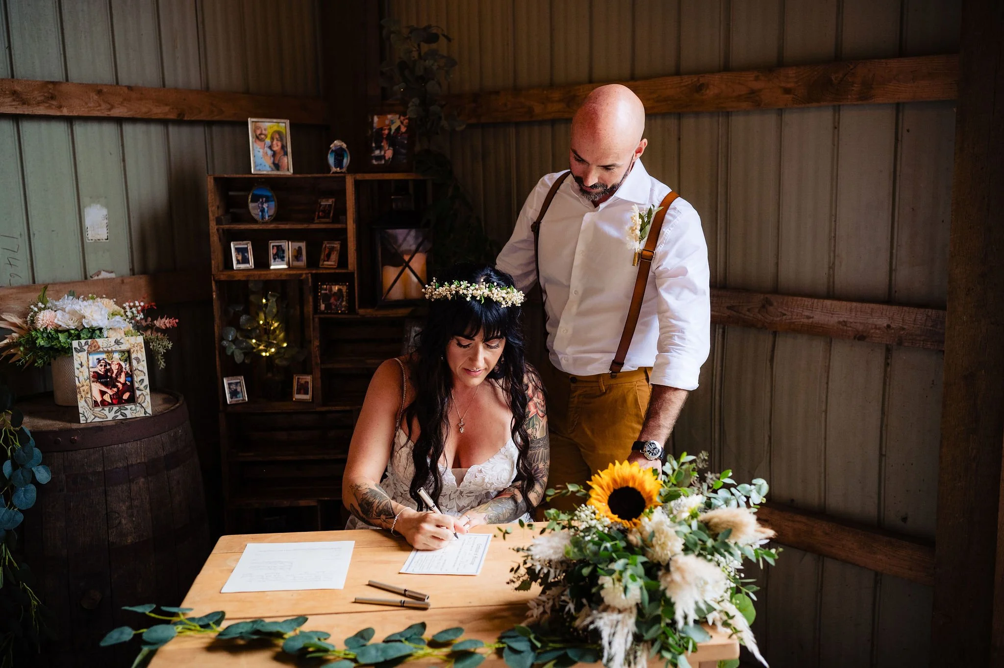 A woman with dark hair, tattoos, and wearing a floral crown signs a document at a wooden table, while a man with a bald head, beard, suspenders, and a white shirt looks on during a wedding ceremony in a rustic setting with framed photos and flowers.