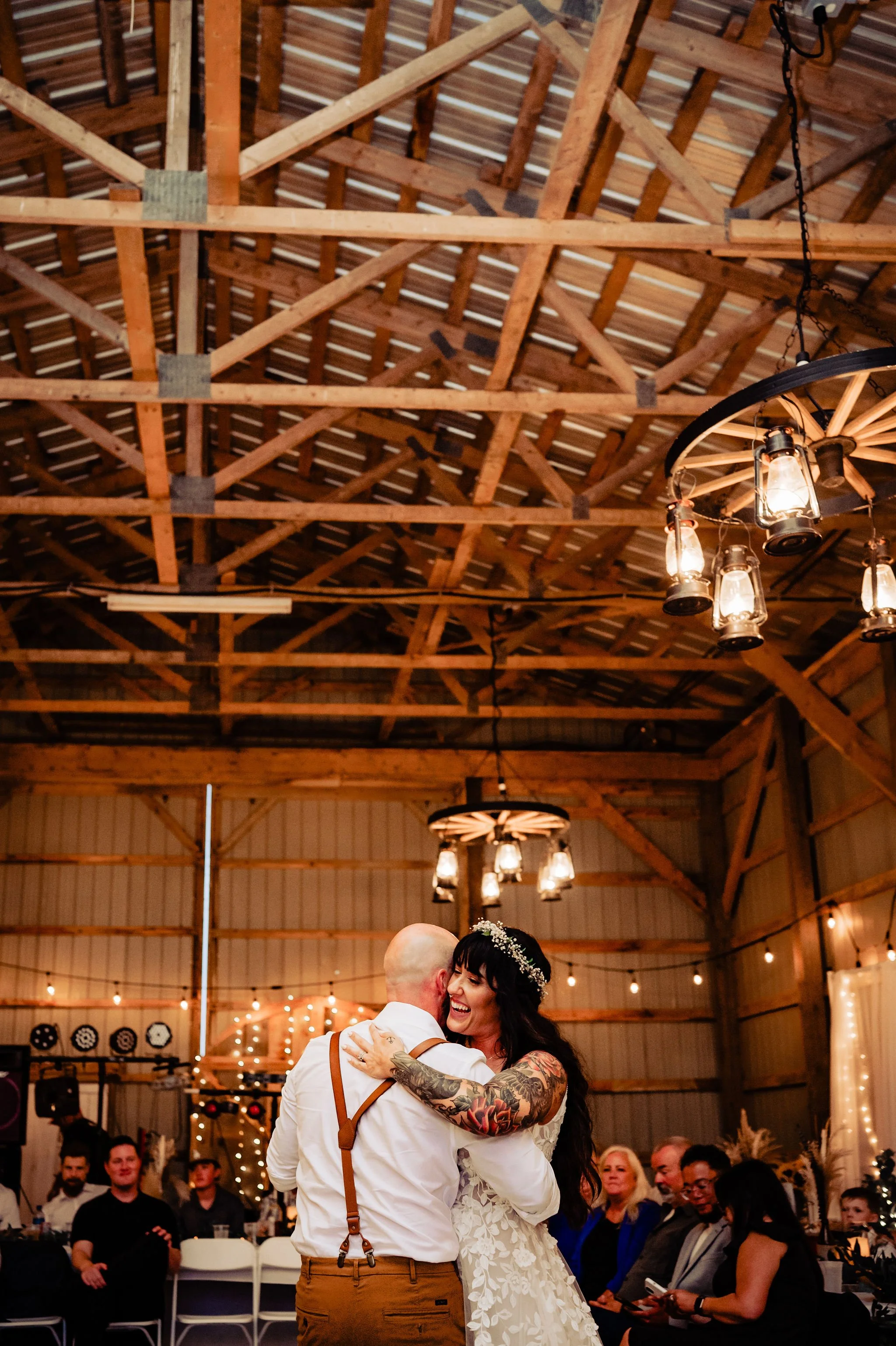 A bride and groom dancing and celebrating at a wedding reception inside a rustic barn, with family and friends seated at tables in the background and decorative lighting overhead.