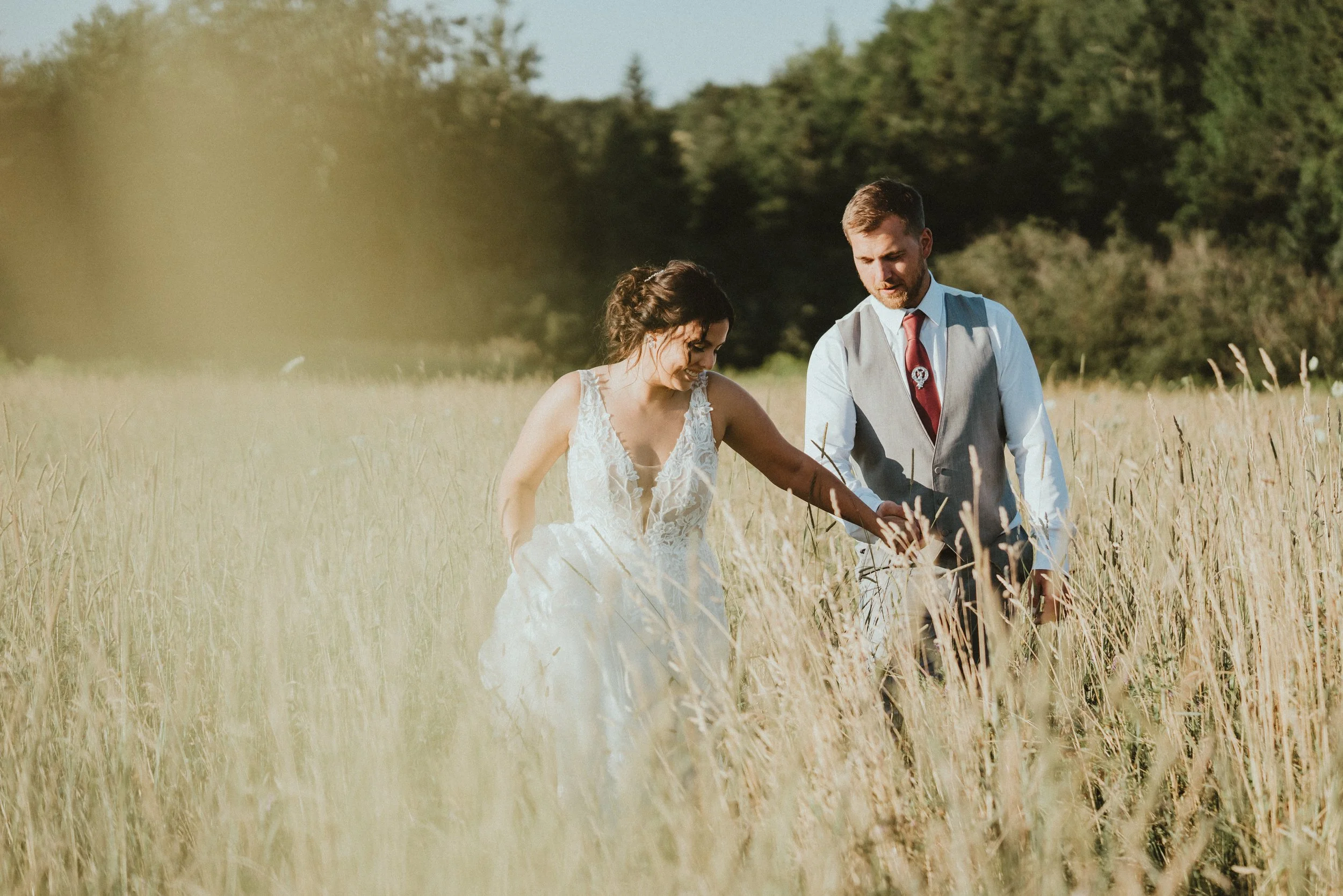 A bride in a white wedding dress and a groom in a vest and tie walking through a field of tall grass on a sunny day.