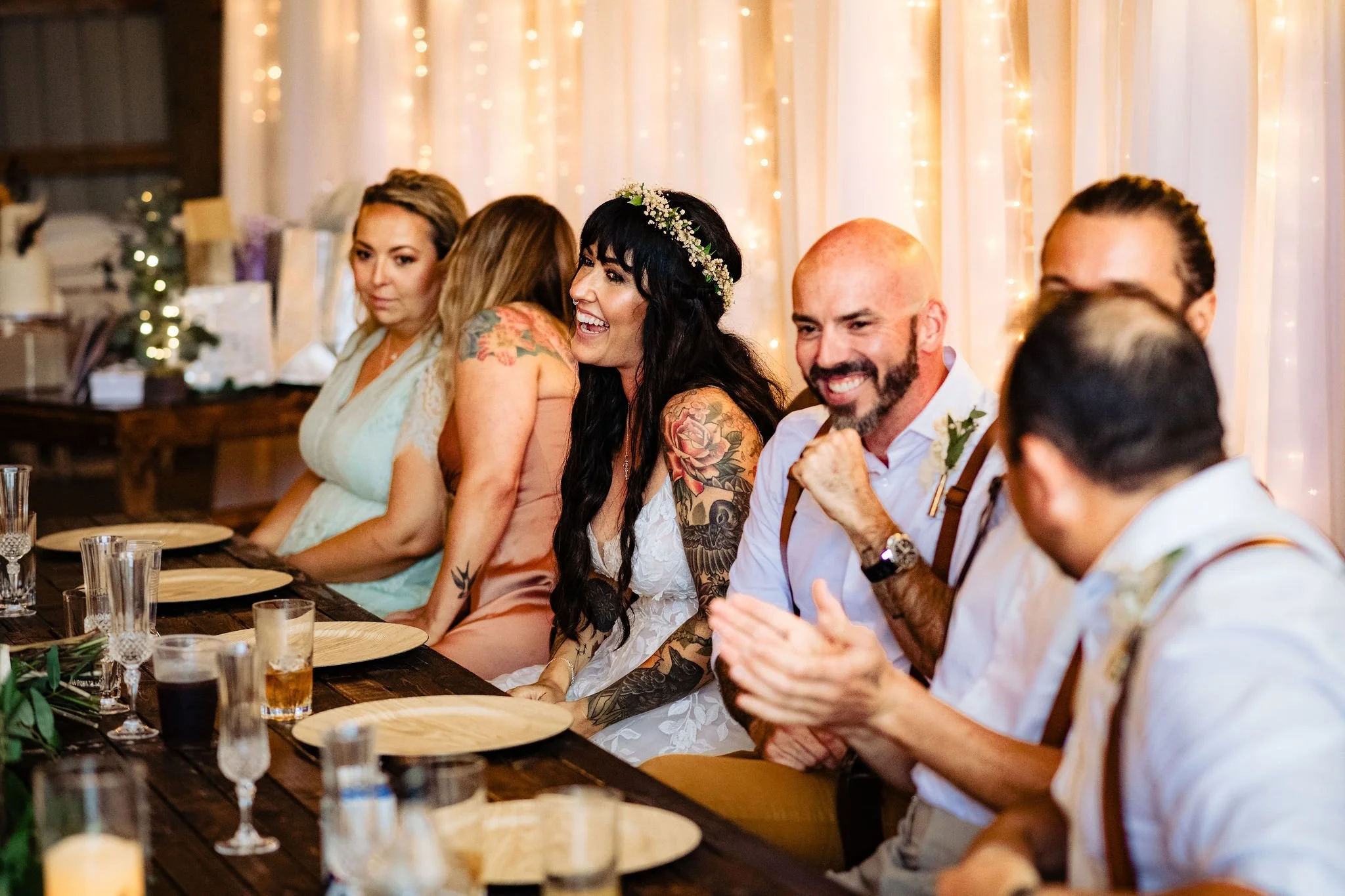 People sitting at a wedding reception, smiling, and clapping, with a decorated backdrop of fairy lights.