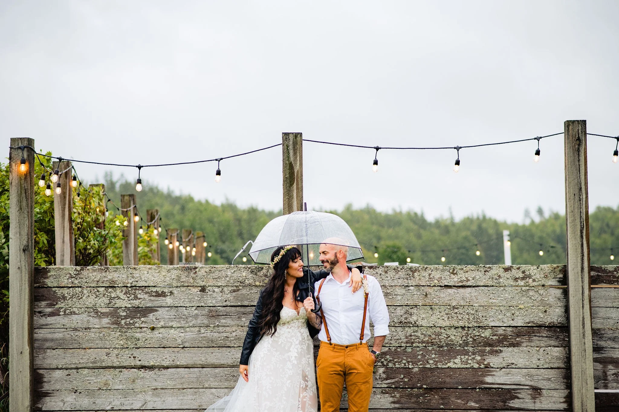 A bride and groom holding a transparent umbrella under a rainy sky, standing close together, smiling at each other, outdoors with string lights and a weathered wooden fence.