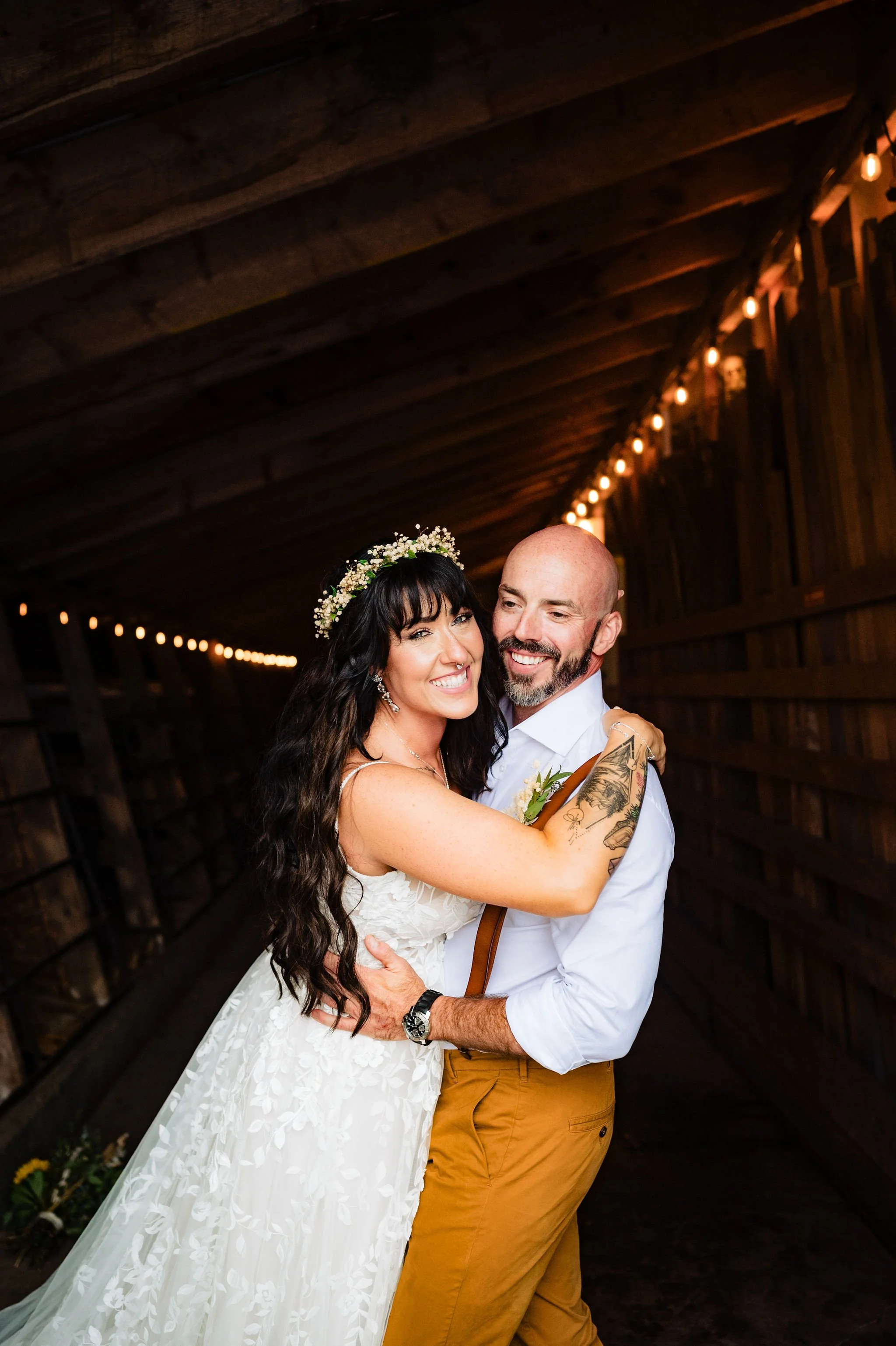 A bride and groom hugging and smiling in a rustic wooden barn with string lights overhead. The bride has long dark hair, a floral crown, and a white lace dress. The groom has a beard, a shaved head, and is wearing a white shirt with yellow suspenders