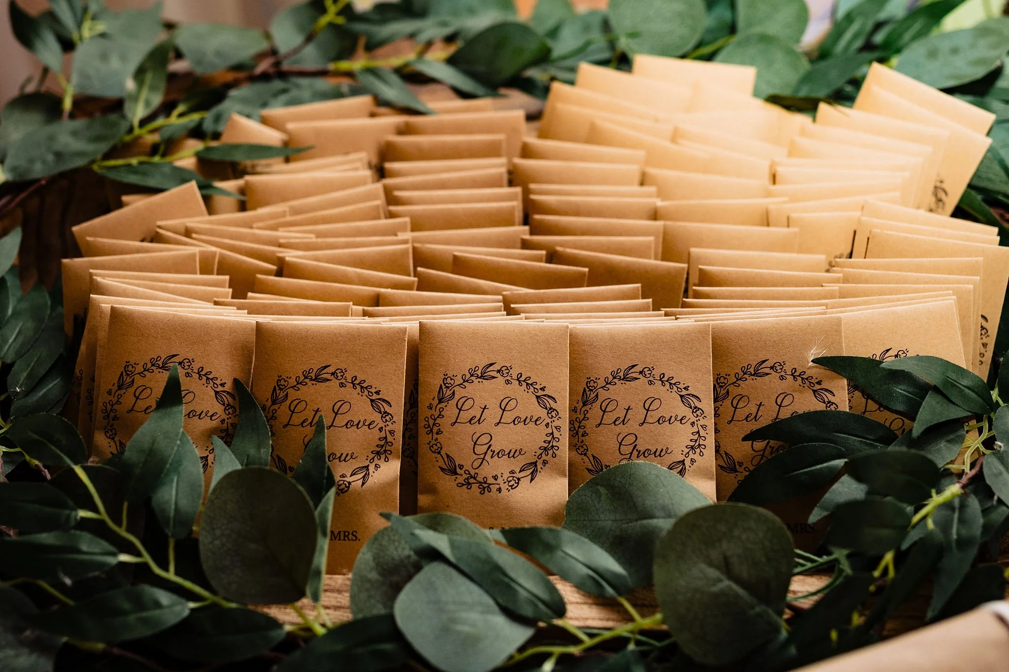 Multiple brown paper bags with the words "Let Love Grow" surrounded by leaf designs, arranged on a table with green leafy branches framing the scene.