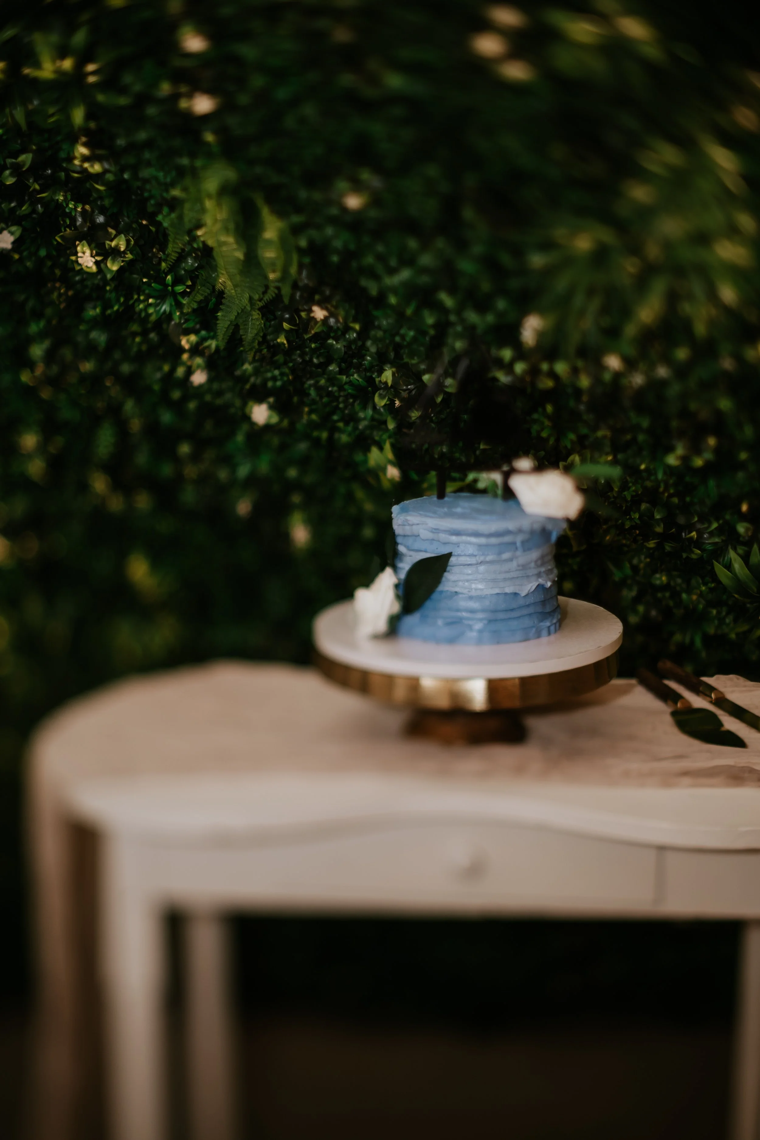 A light blue frosted cake with black topper and white flowers on a wooden table in front of a green leafy backdrop.