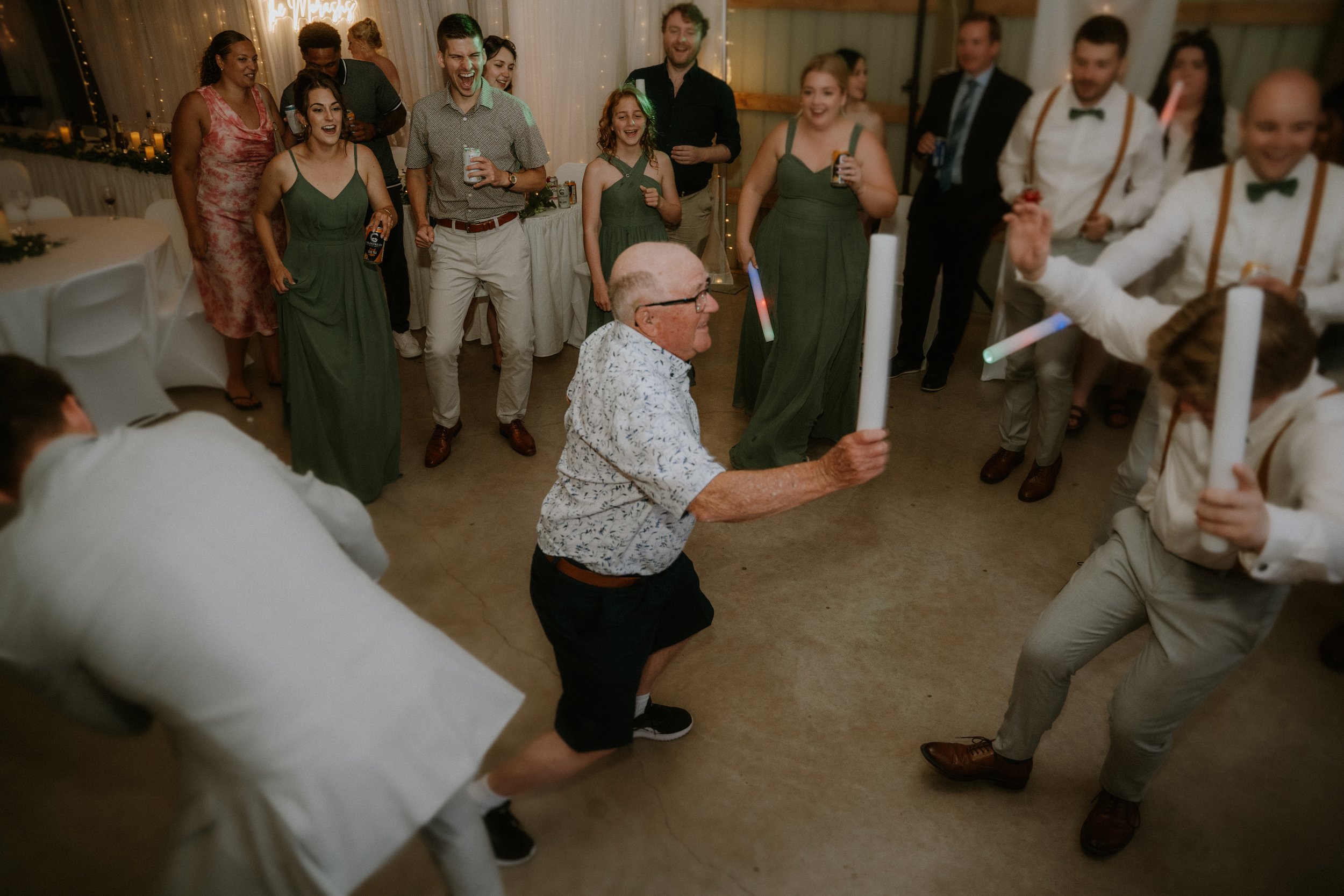 Elderly man and young man dancing with light-up sticks at wedding reception, surrounded by guests