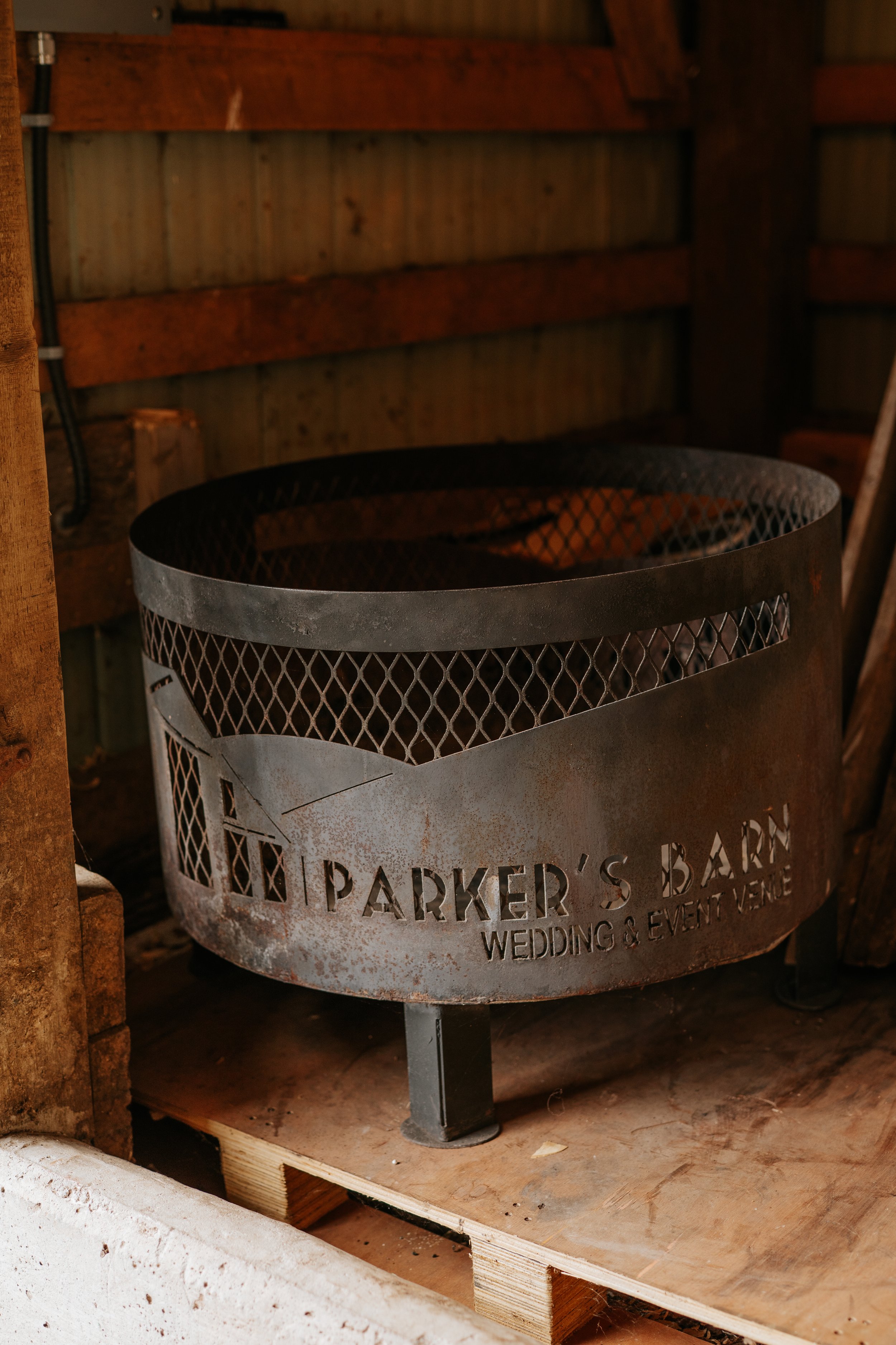 Round metal fire pit with cutout house graphic and the text 'Parker's Barn Wedding & Event Venue', placed on a wooden pallet in a rustic wooden shed.