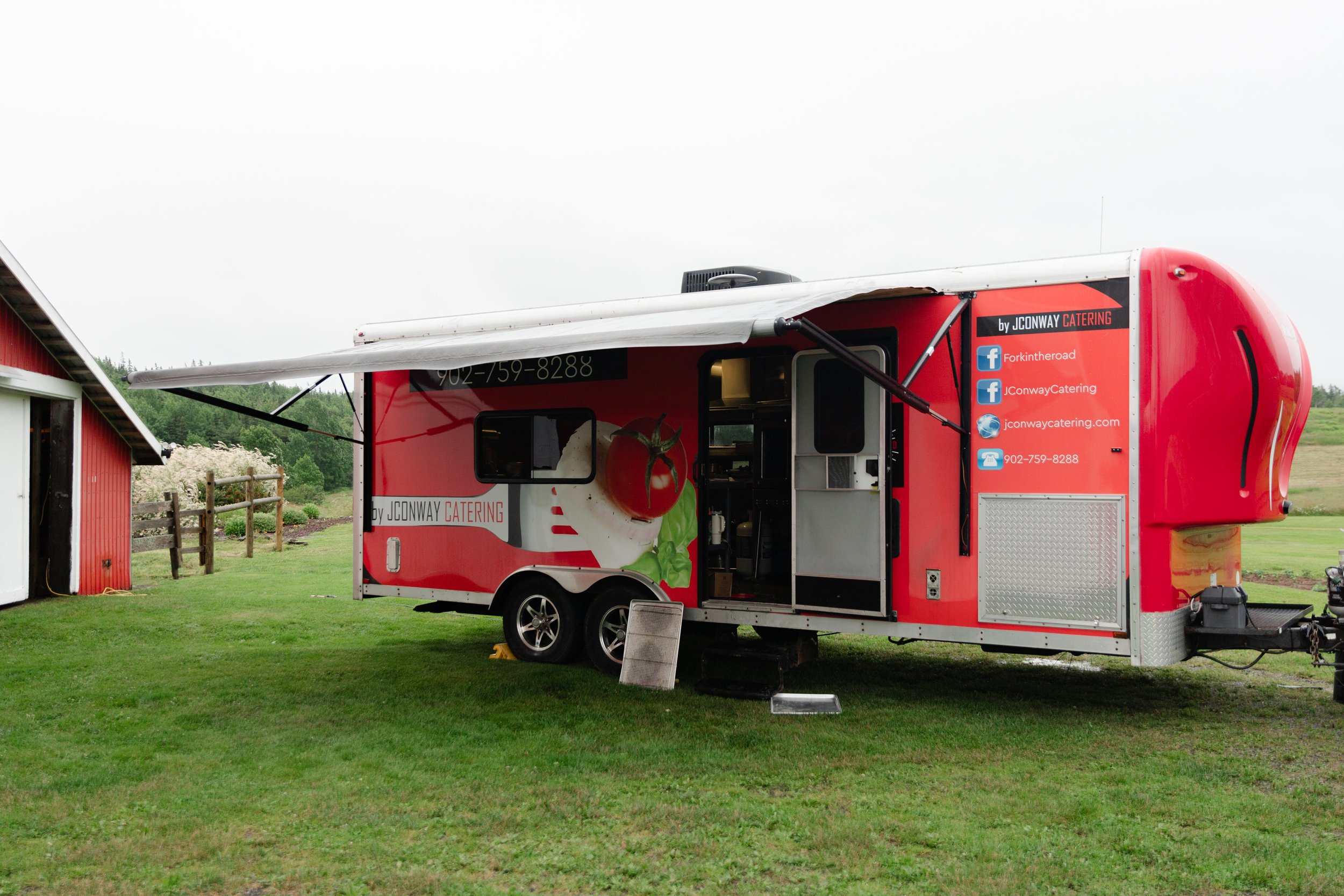 Red food truck with graphics of a tomato and lettuce, parked on a grassy field near a red barn and trees, with the side window open and advertising for JCONWAY CATERING.