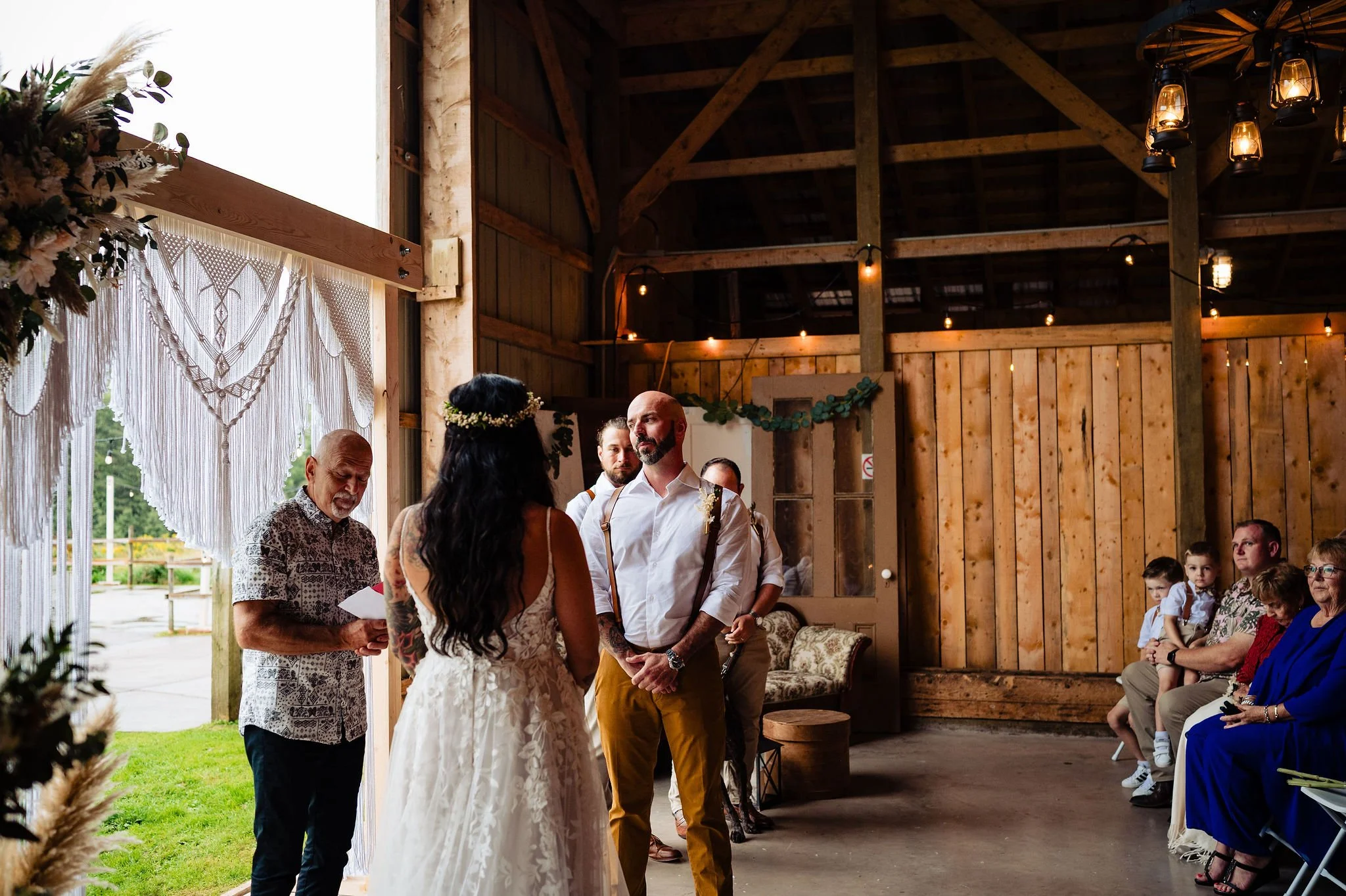 A wedding ceremony taking place in a rustic wooden barn, with a bride and groom facing each other, surrounded by seated guests and an officiant reading from a paper.