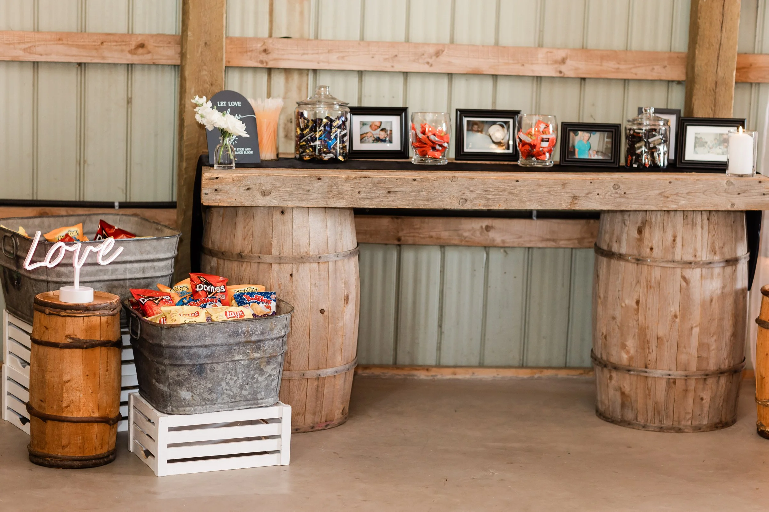 Rustic setup with a wooden table on large wine barrel bases, framed photographs, jars filled with candies, a candle, and snack baskets including bags of chips.