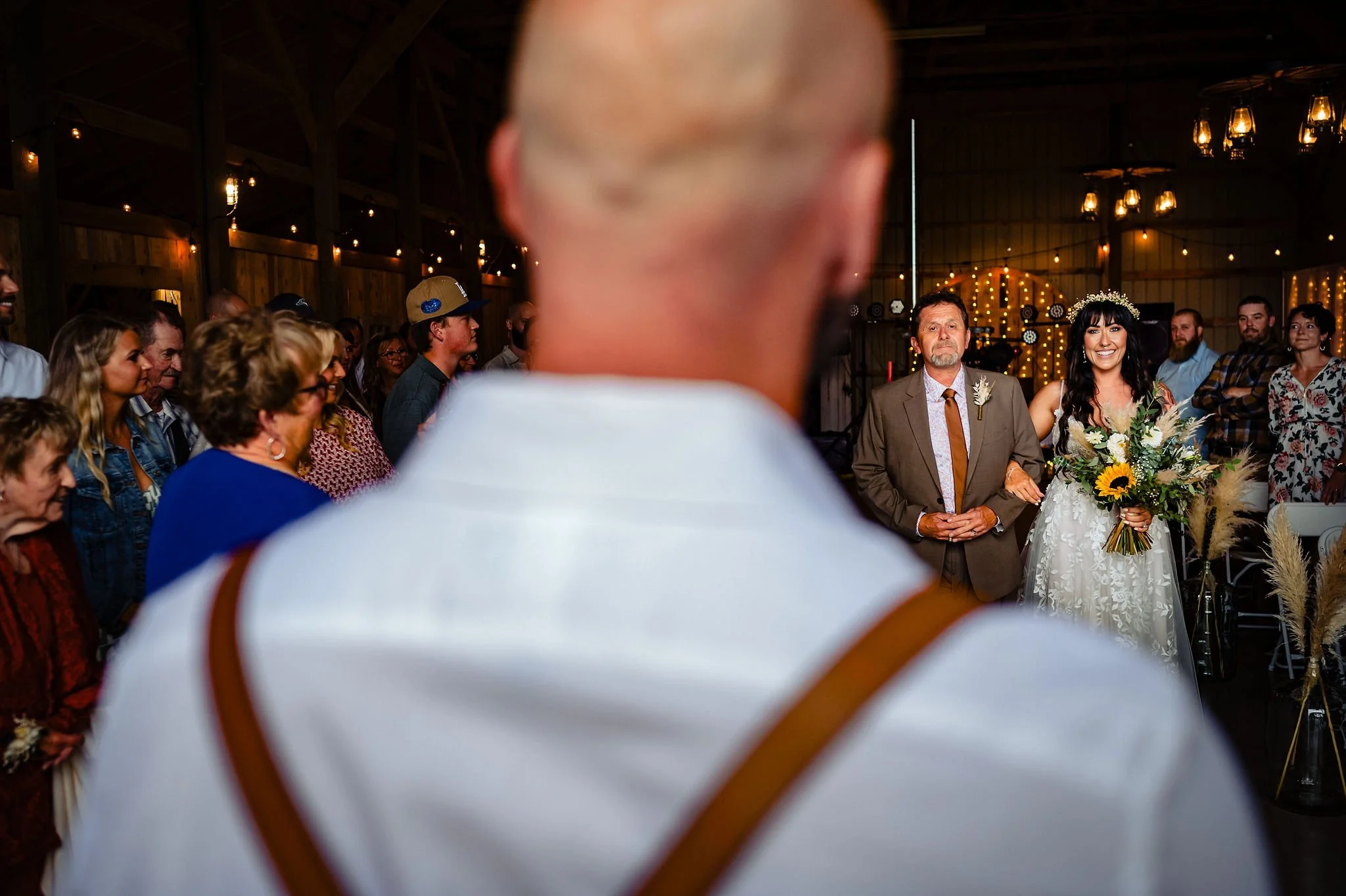 A bride smiling with a bouquet of flowers, accompanied by a man in a suit, at a wedding ceremony inside a barn decorated with string lights.