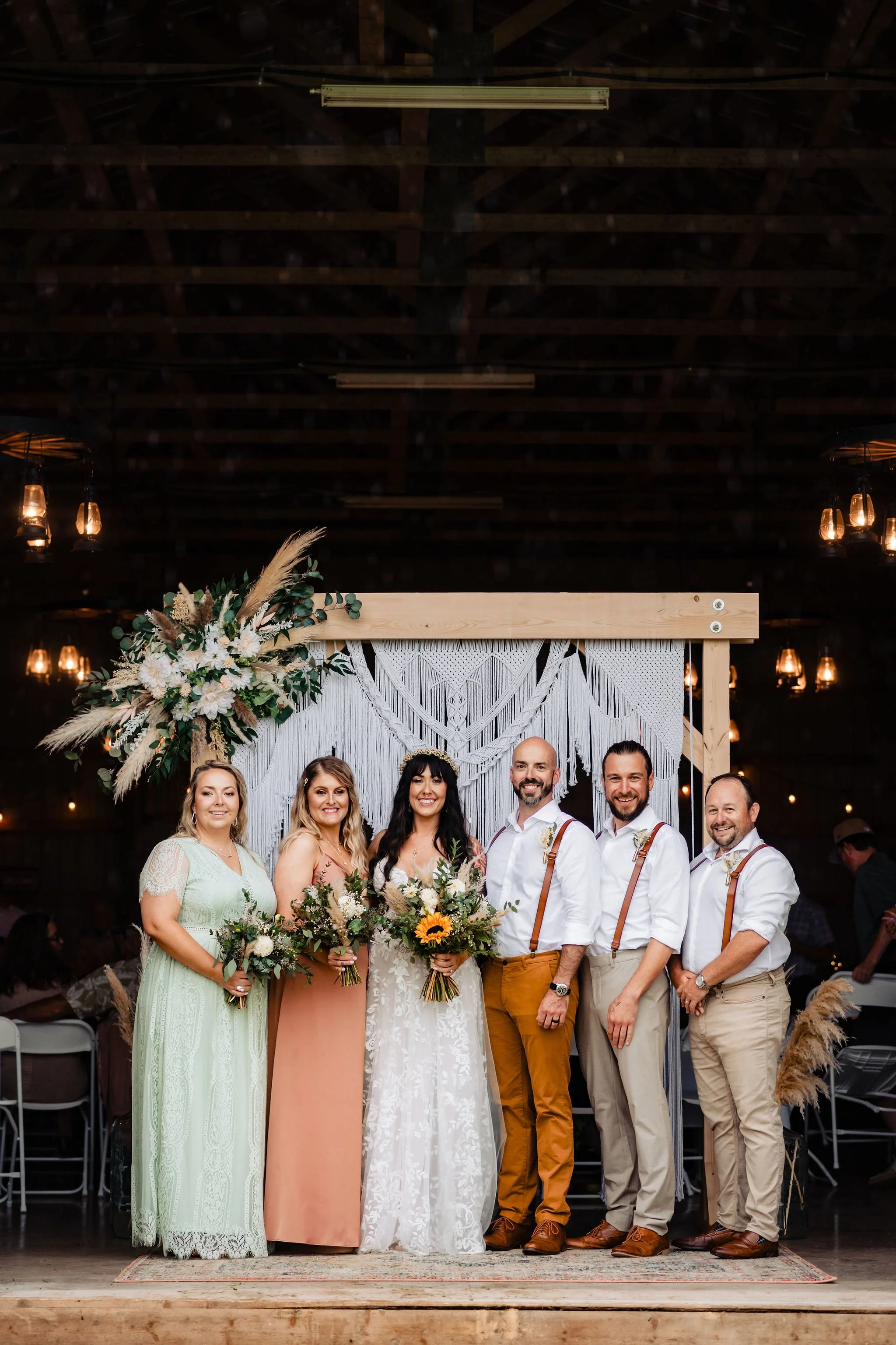 A wedding party standing together in front of a decorated backdrop, holding bouquets, in a rustic barn setting with hanging lantern lights.