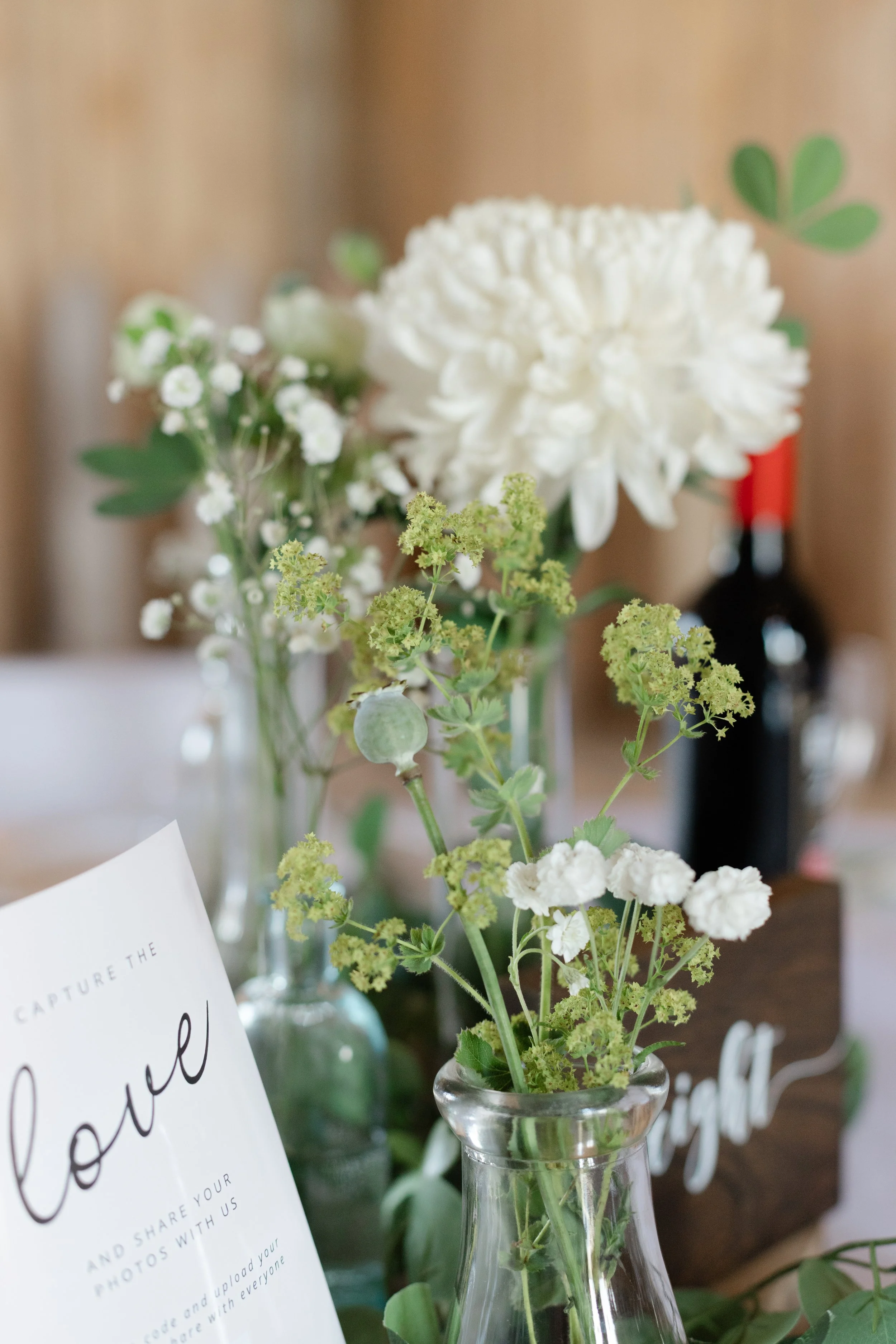 Close-up of small white and green flowers in glass vases, with a sign mentioning sharing photos and a red wine bottle in the background.