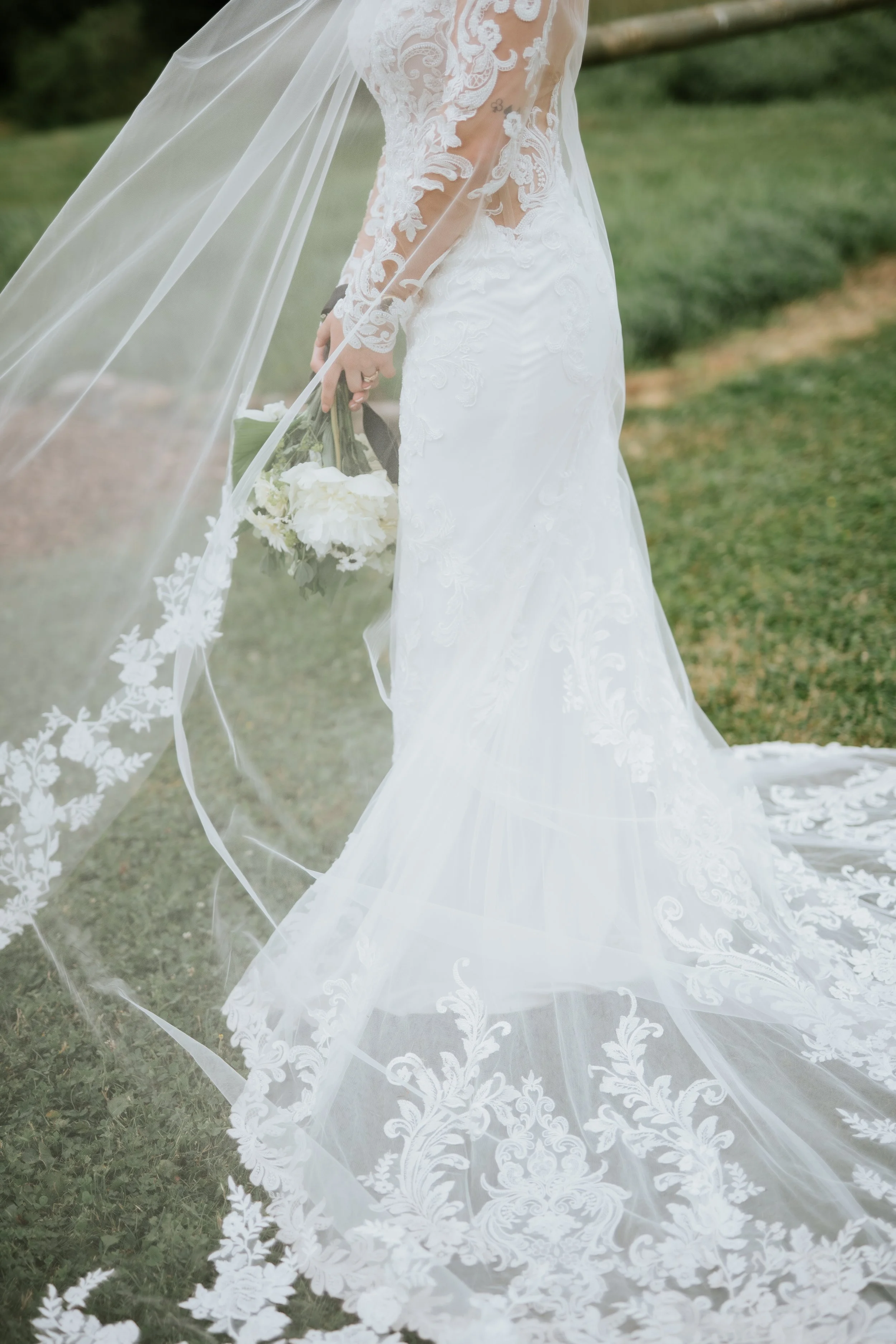 Close-up of a bride in a white lace wedding dress holding a bouquet of white flowers, standing on grass outdoors.