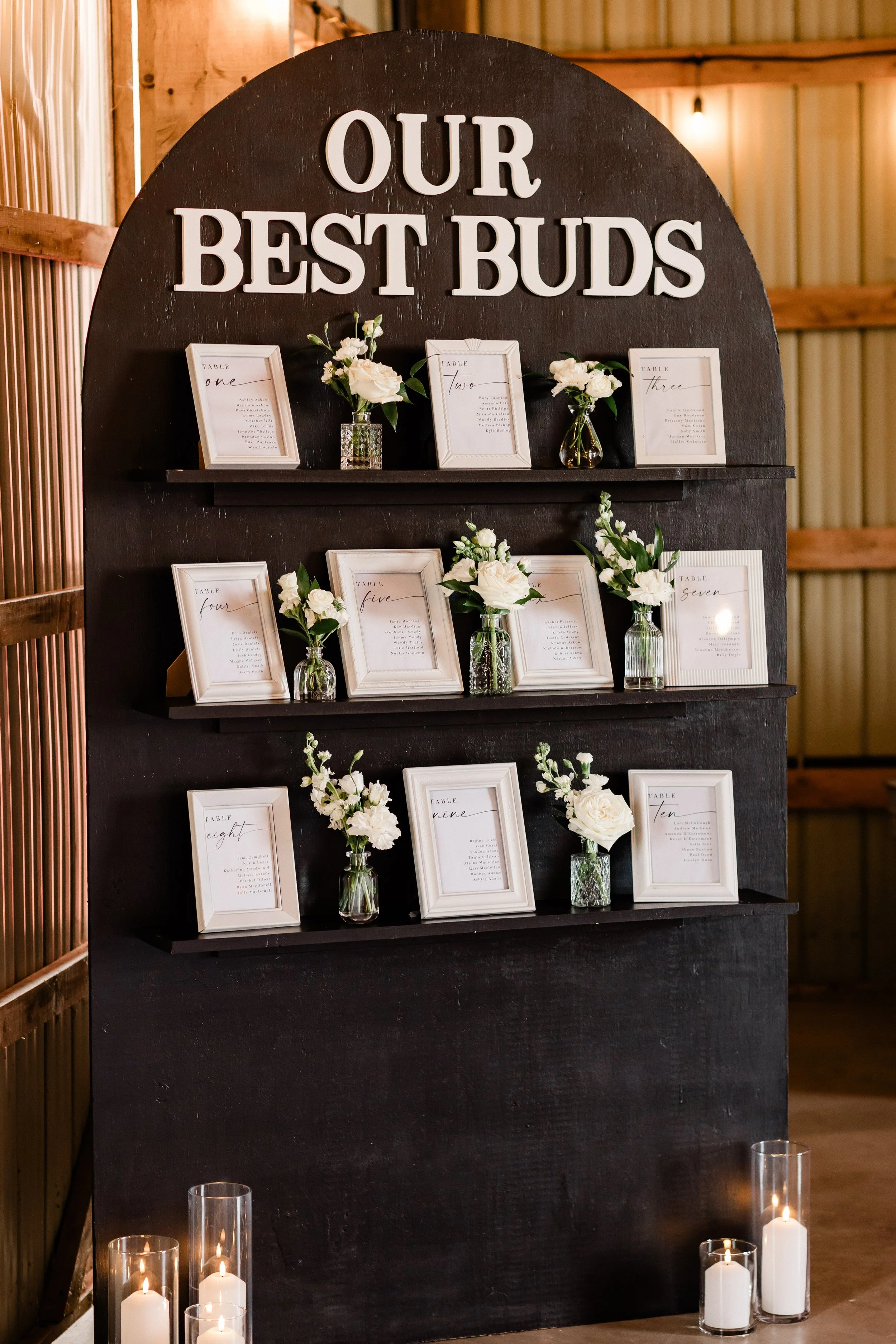 Table seating arrangement display with framed signs and floral arrangements on black shelves, titled 'Our Best Buds' at a rustic wedding or event.