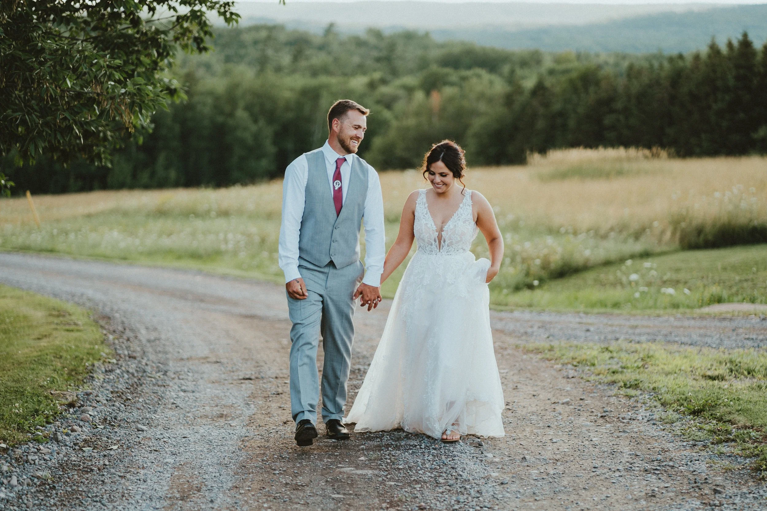 A newlywed couple walking hand in hand on a dirt road in a countryside setting, smiling and looking at each other. The bride is in a white lace wedding gown with pockets, and the groom is in a light gray suit with a red tie. Surrounding them are gree