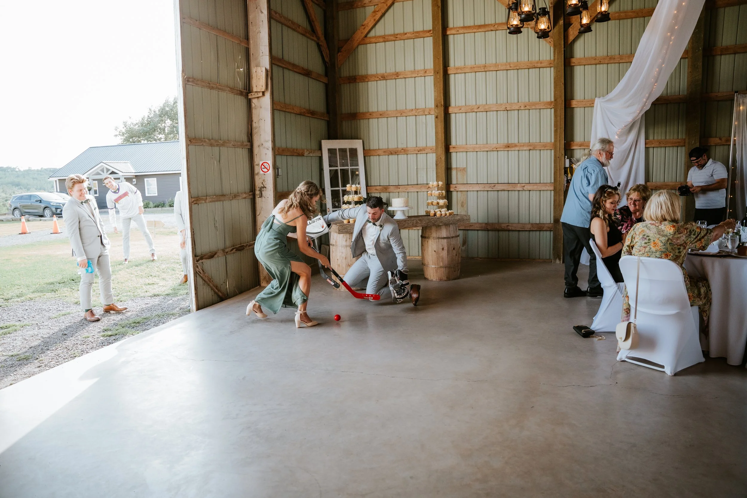 People playing hockey inside a rustic barn at a celebration or wedding, with some guests sitting at a round table and others outside near a parking lot.