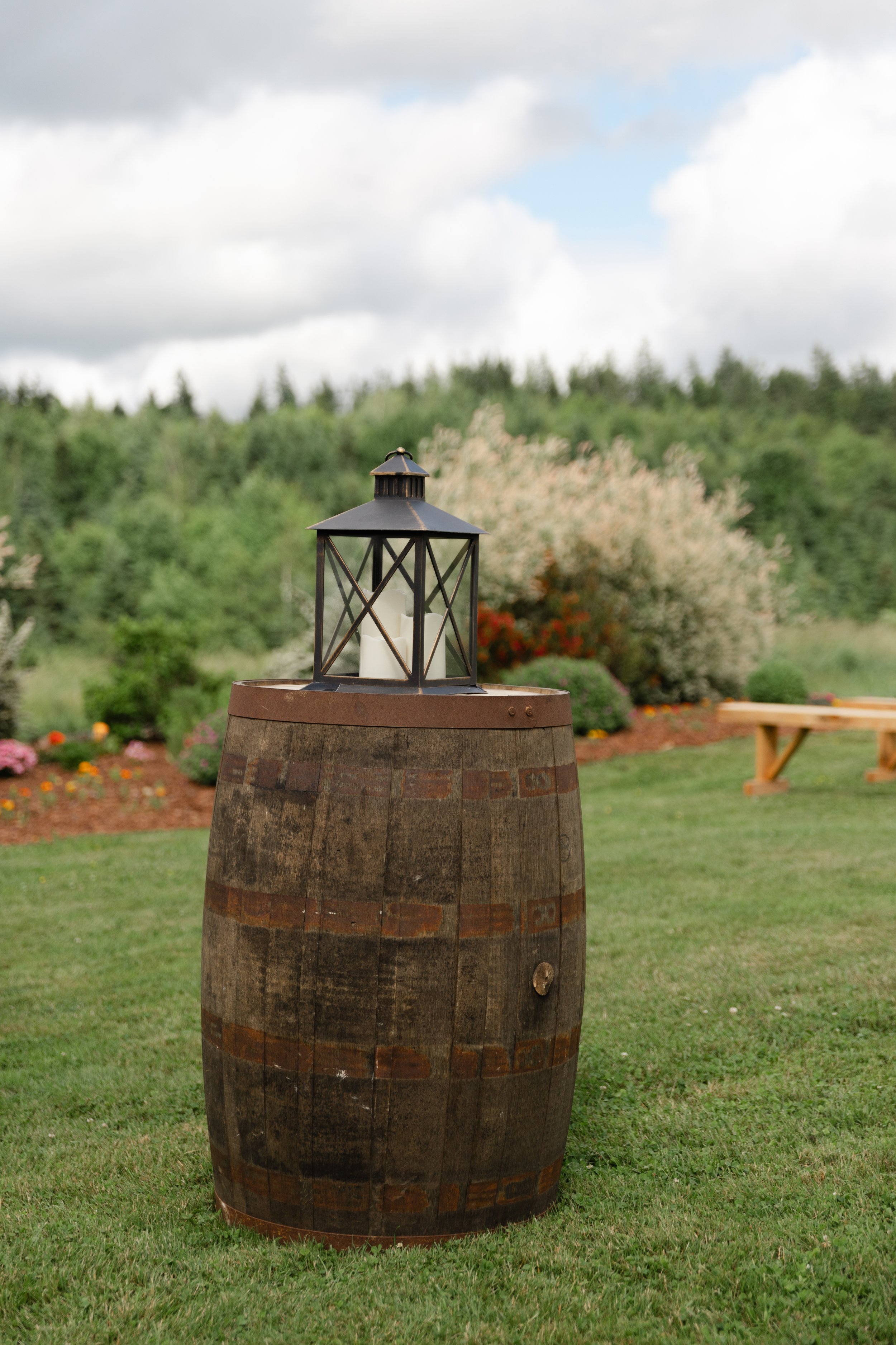 A wooden barrel with a metal lantern on top, set on a grassy lawn in a garden with colorful flowers and green trees in the background under a cloudy sky.