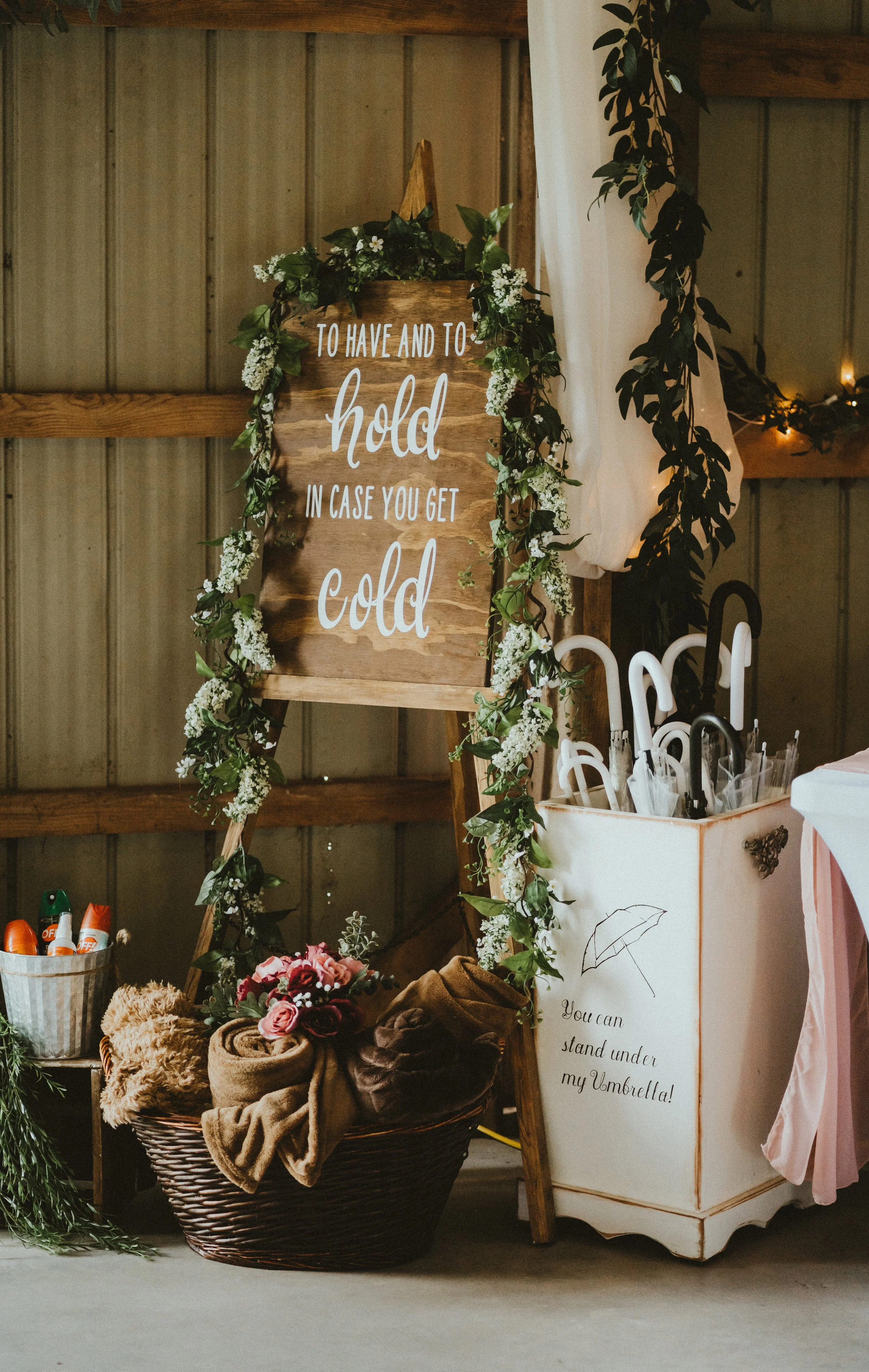 A decorative stand with a wooden sign that reads 'To have and to hold in case you get cold,' surrounded by greenery and white flowers. There are umbrellas in a white container nearby, with a sign that says 'You can stand under my umbrella!'. A basket