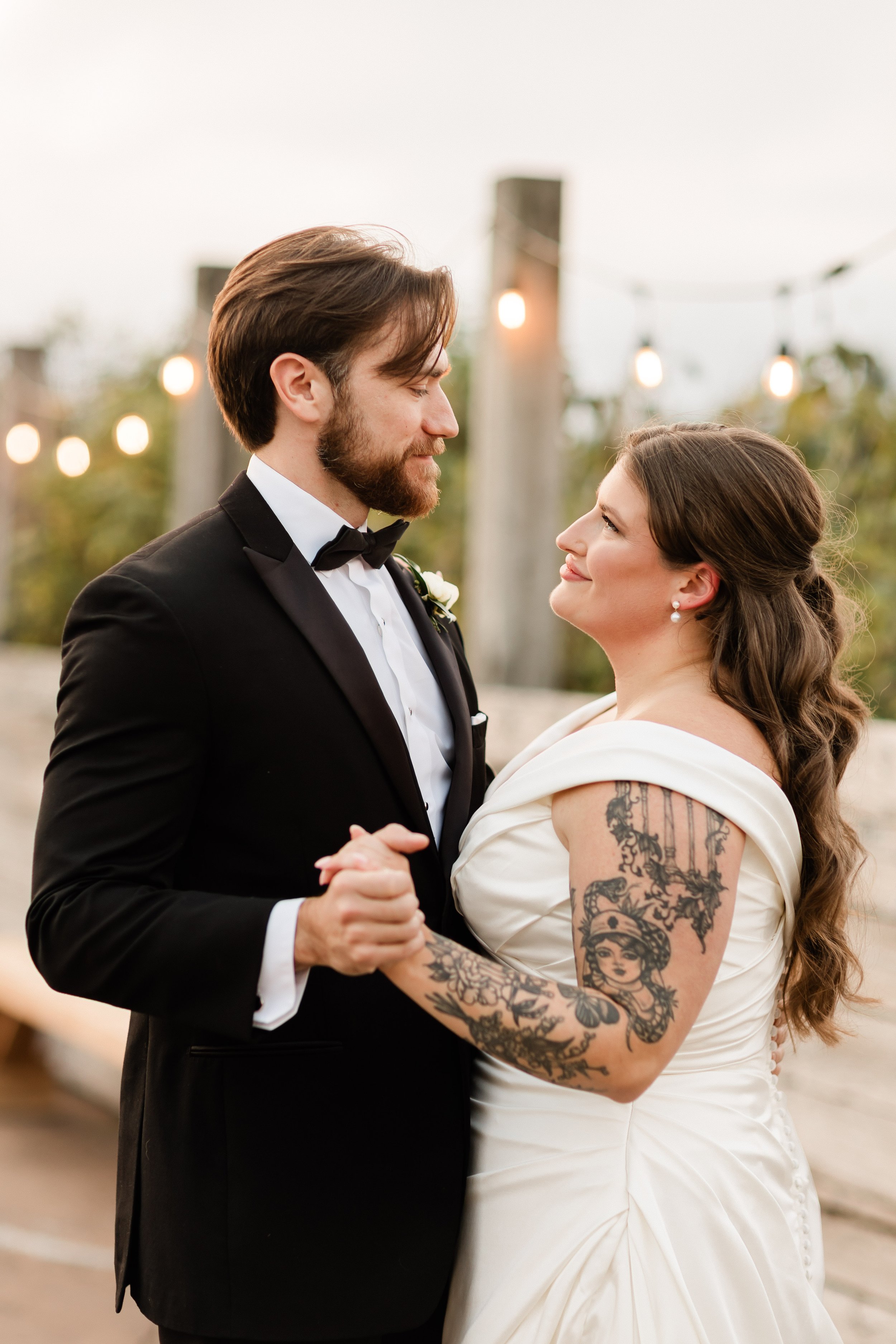 A couple in wedding attire dancing outdoors with string lights in the background.