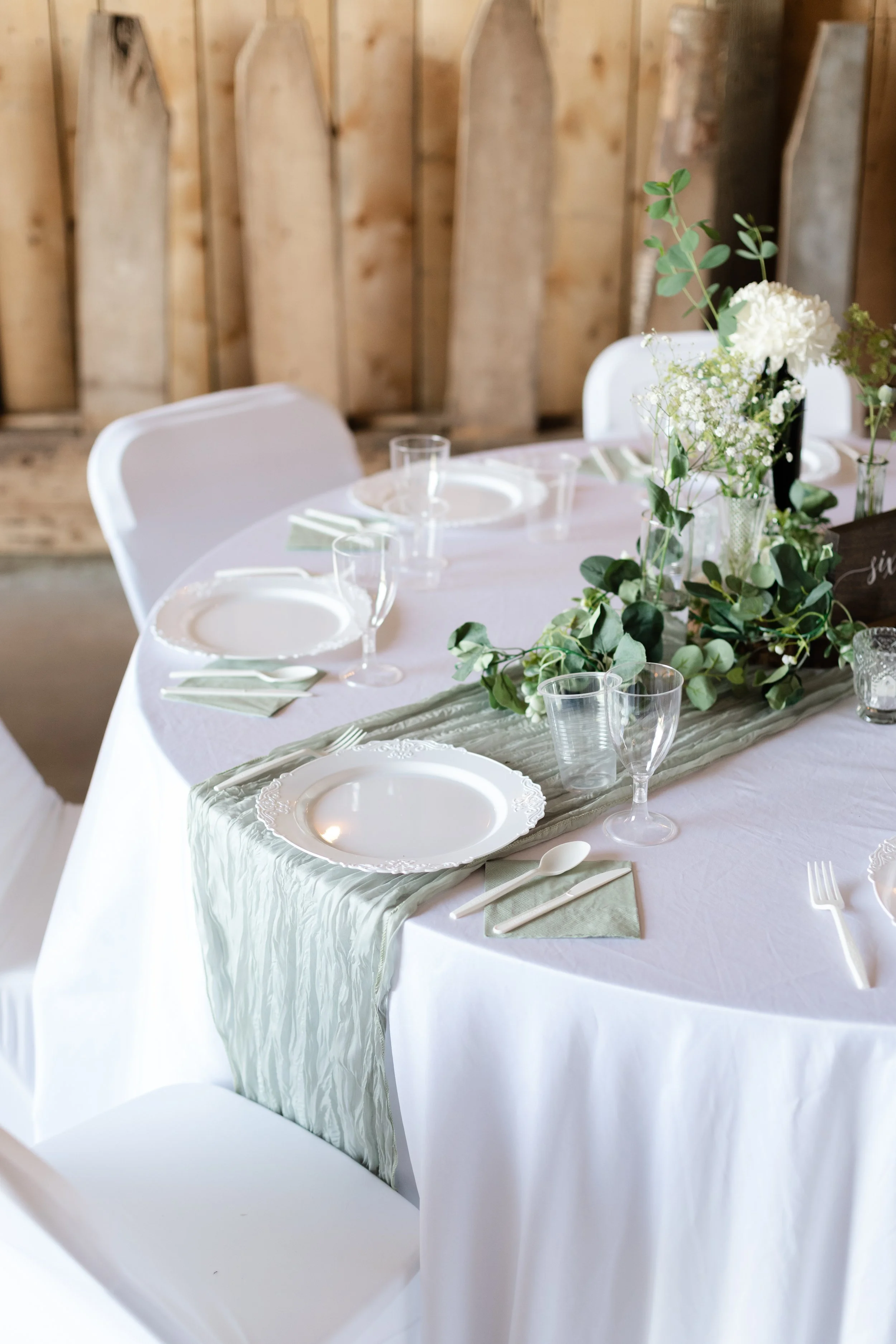 Wedding reception table with white tablecloth, green runners, white plates, green napkins, clear glasses, floral centerpieces with white flowers and greenery, and white chairs in a rustic setting.
