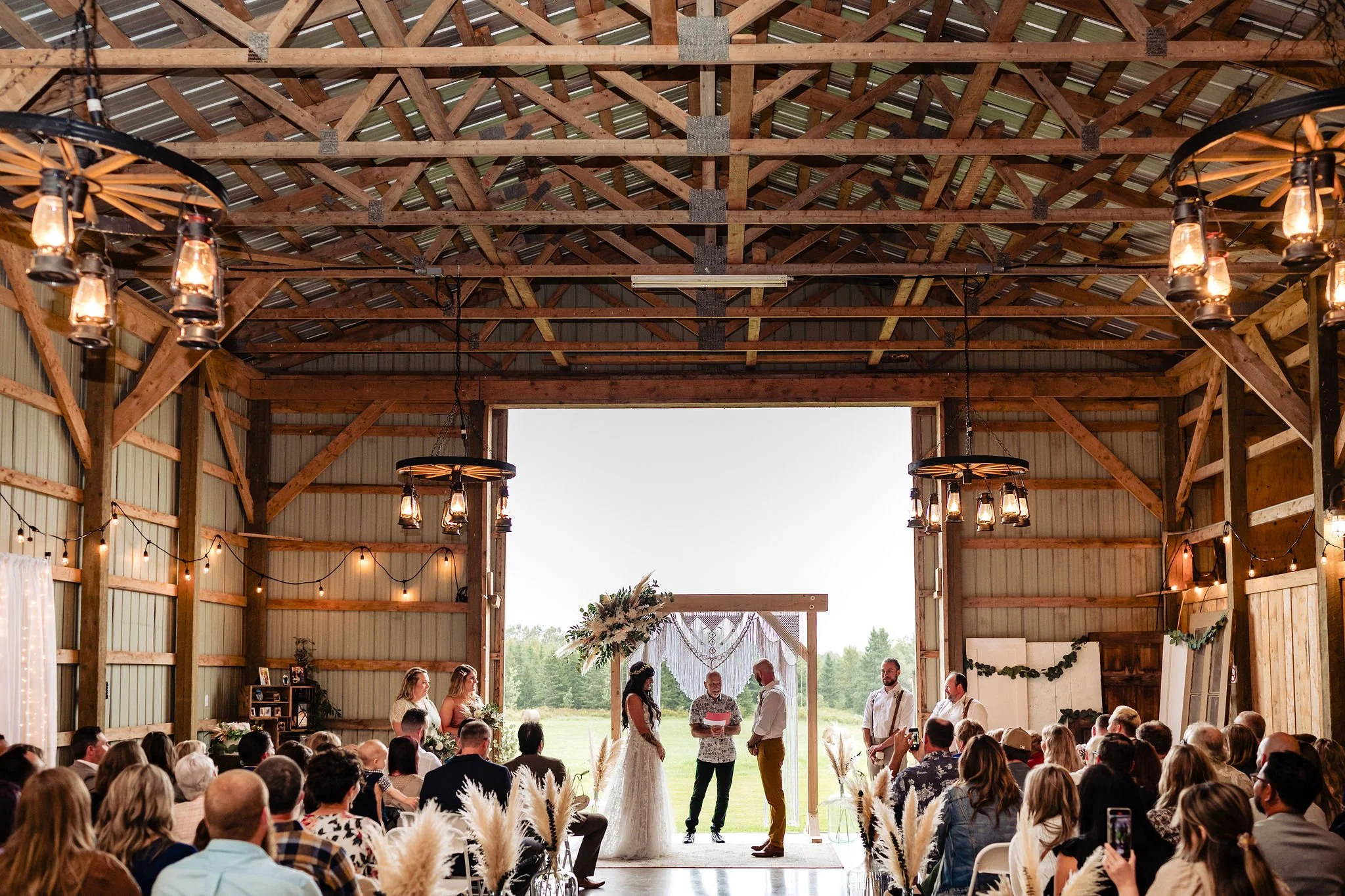 A wedding ceremony inside a barn with wooden beams, string lights, and a large open doorway showing a green landscape outside. The bride and groom stand in front of a small altar, with an officiant reading from a paper. Bridesmaids and groomsmen are 