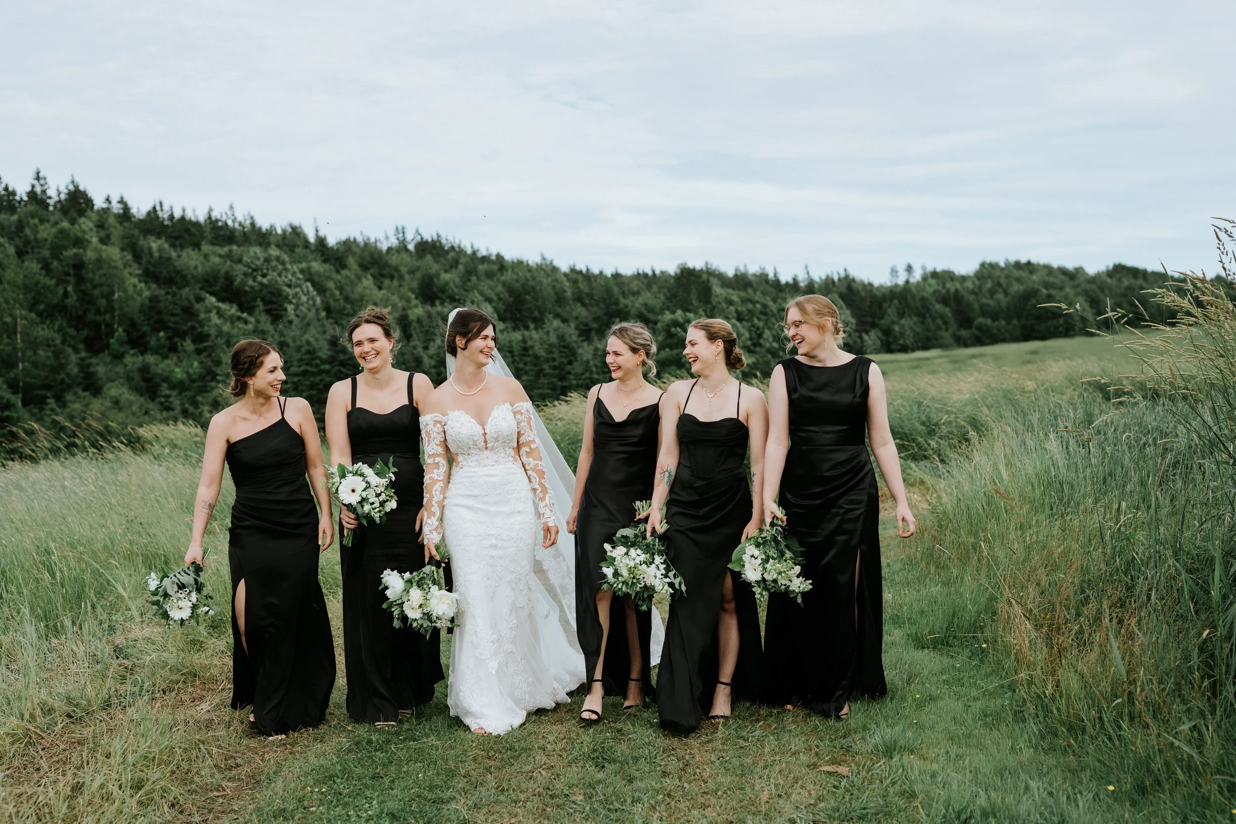 A bride and five bridesmaids walk together in a grassy field, all smiling and holding bouquets of white flowers, with green trees and a cloudy sky in the background.