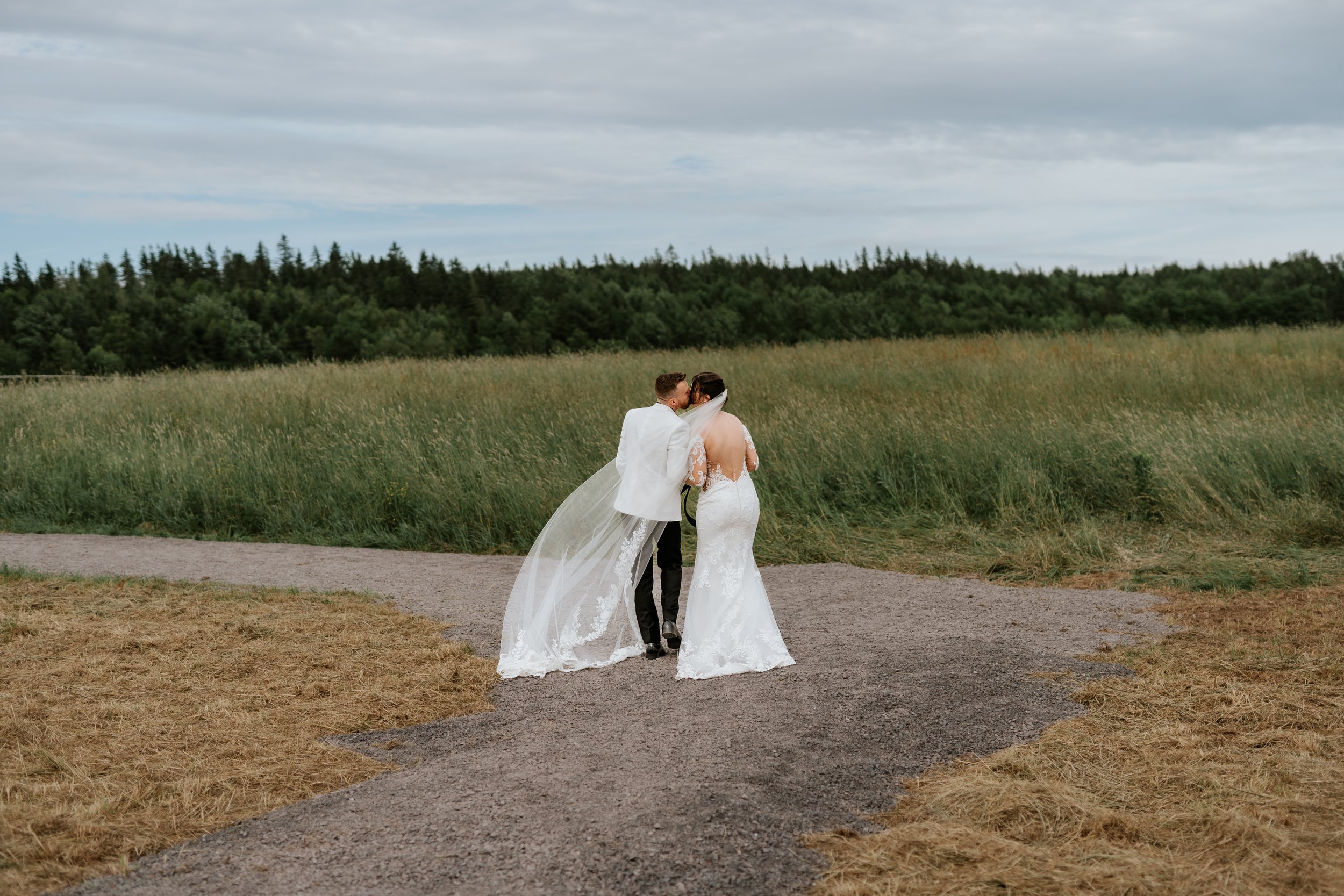 A bride and groom walking on a dirt path in a field, with the groom leaning in to kiss the bride's cheek. The bride is wearing a white lace wedding gown with long sleeves and a veil, and the groom is dressed in a white jacket and black pants. The fie