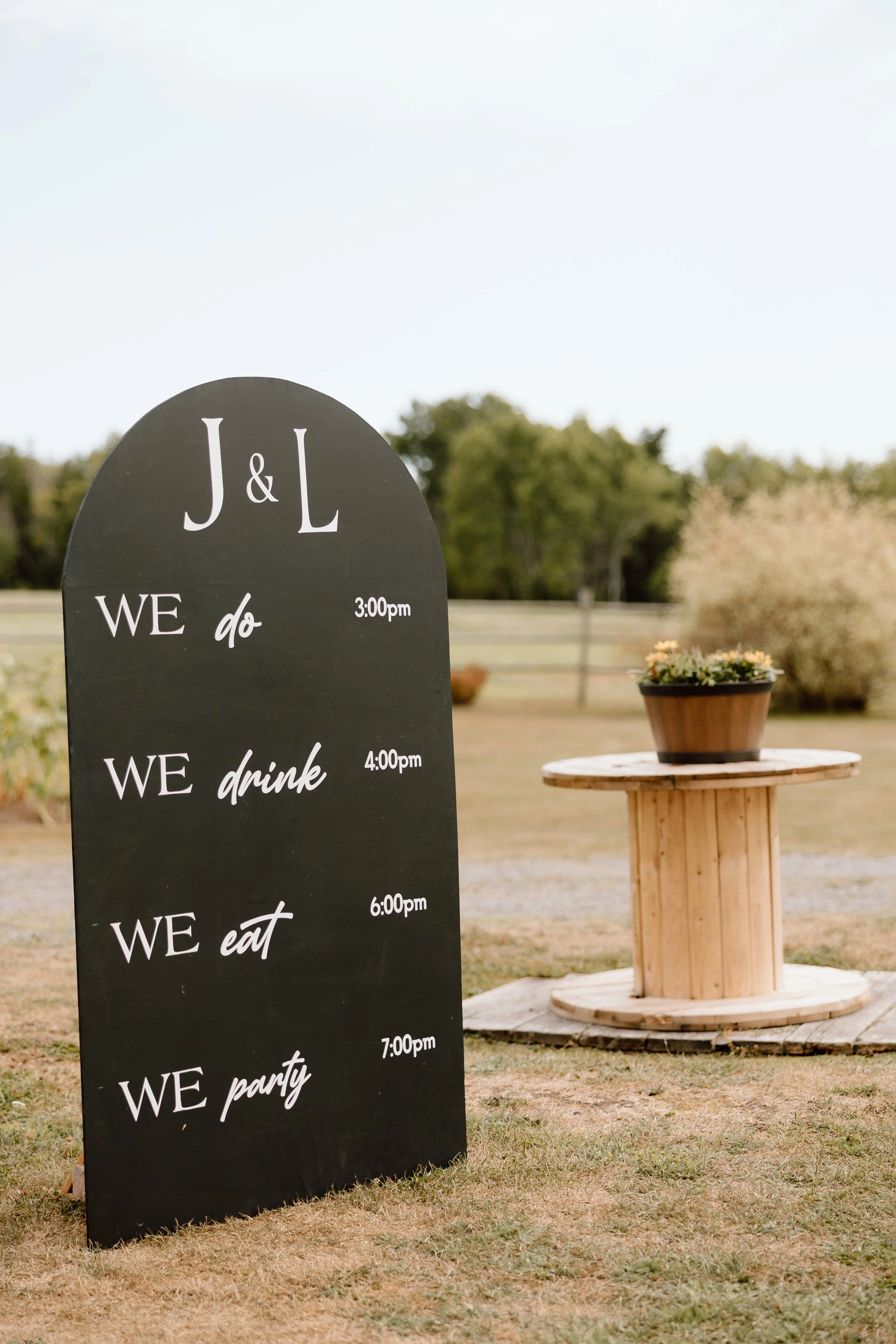 Outdoor event schedule sign listing activities from 3:00 pm to 7:00 pm, including with times next to each, next to a wooden table with a potted plant, in a grassy area with trees in the background.