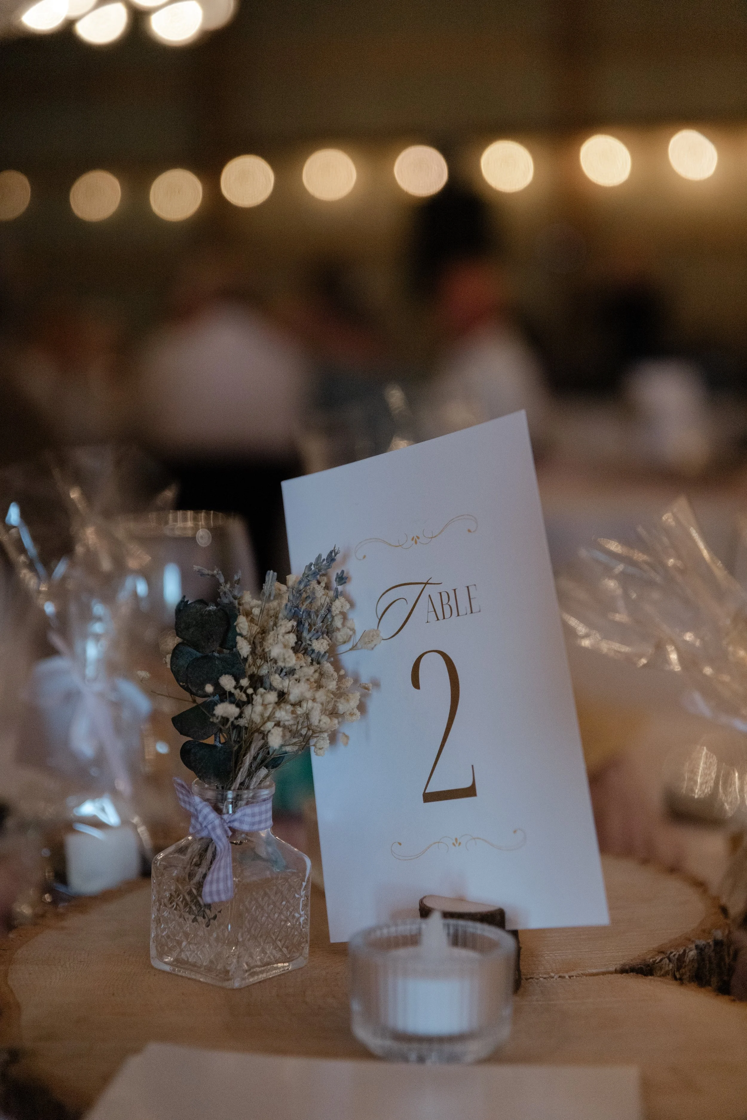 Wedding reception table with a table number 2 card, small dried flower arrangement in a glass vase, and a tea light candle in a glass holder, with blurred guests and warm lighting in the background.