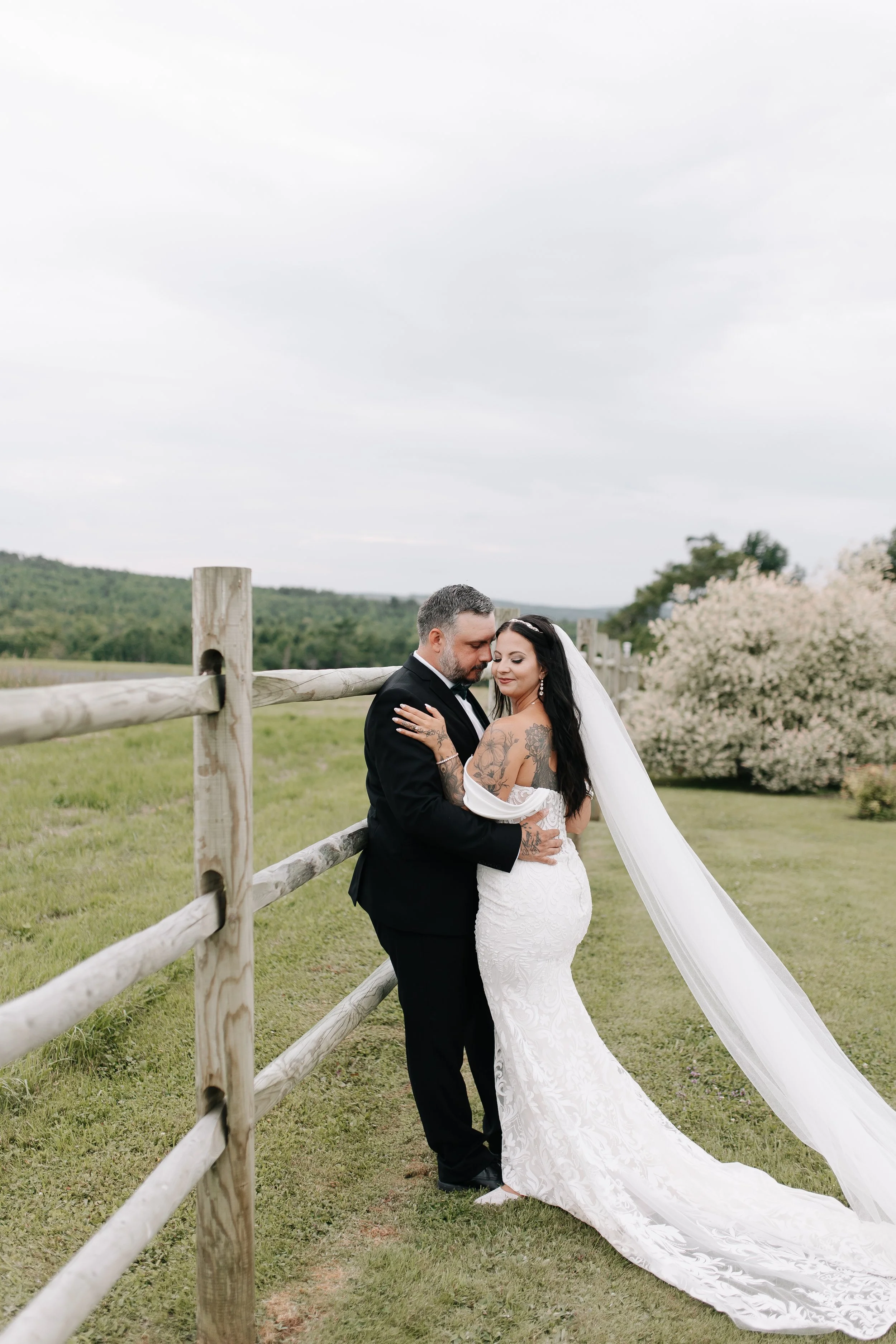 Bride and groom embracing outdoors in a grassy field with a wooden fence, trees, and overcast sky.