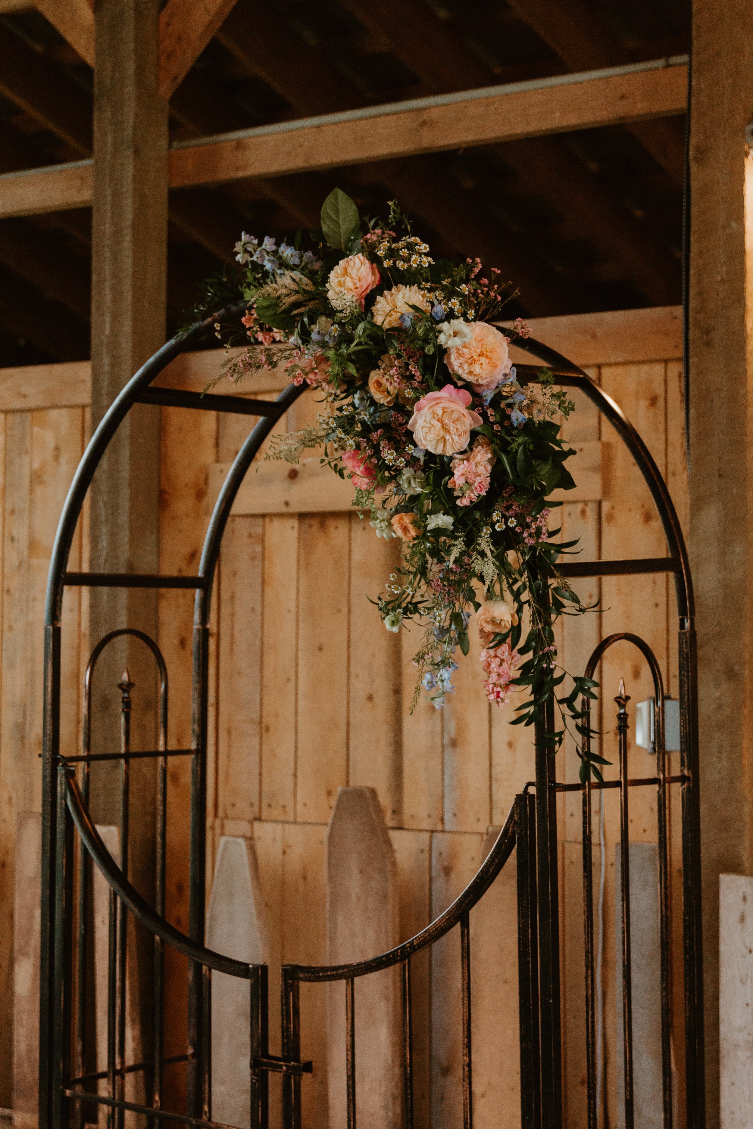 A black metal wedding arch decorated with pink and peach flowers and greenery, set against a wooden barn interior.