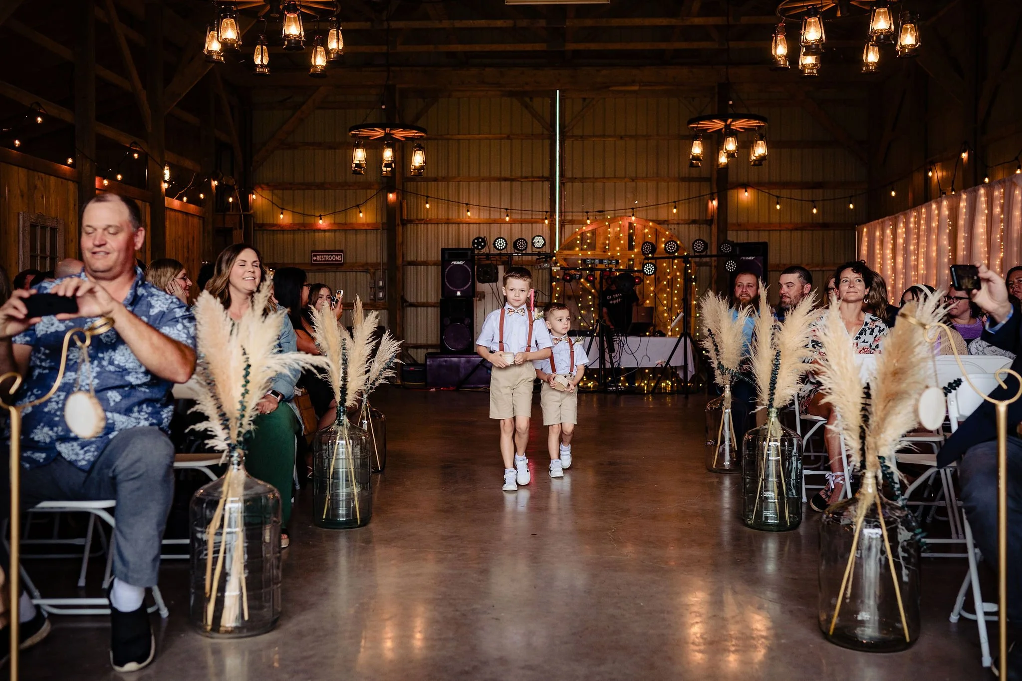 Children walking down the aisle at a wedding ceremony in a rustic barn decorated with string lights and pampas grass in vases, with guests seated on either side taking photos and watching.