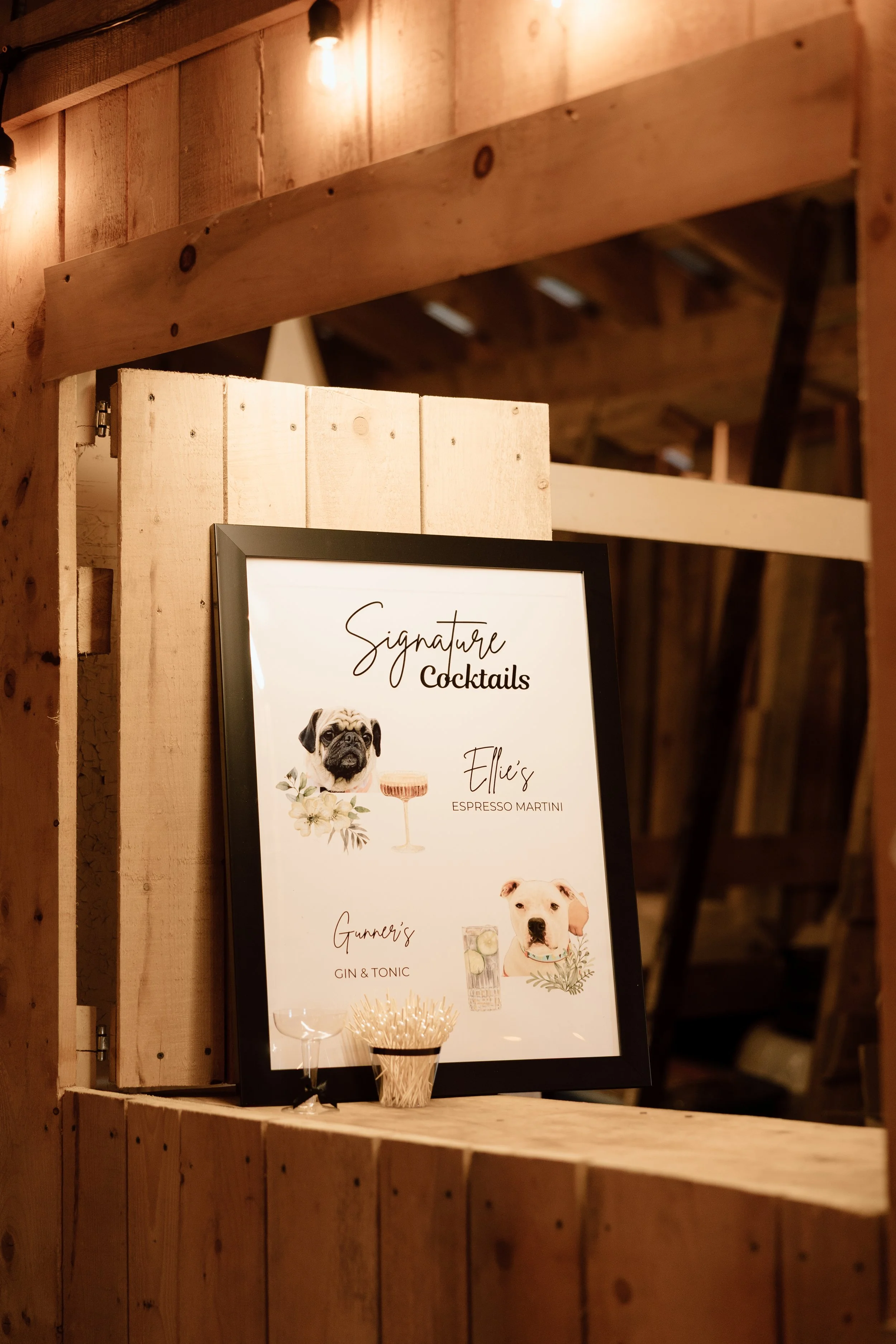 A framed sign displays a menu of signature cocktails at a rustic venue, featuring illustrations of a pug and a dog with a list of cocktails including Ellie’s Espresso Martini and Gunner’s Gin & Tonic, surrounded by a small container of toothpicks and
