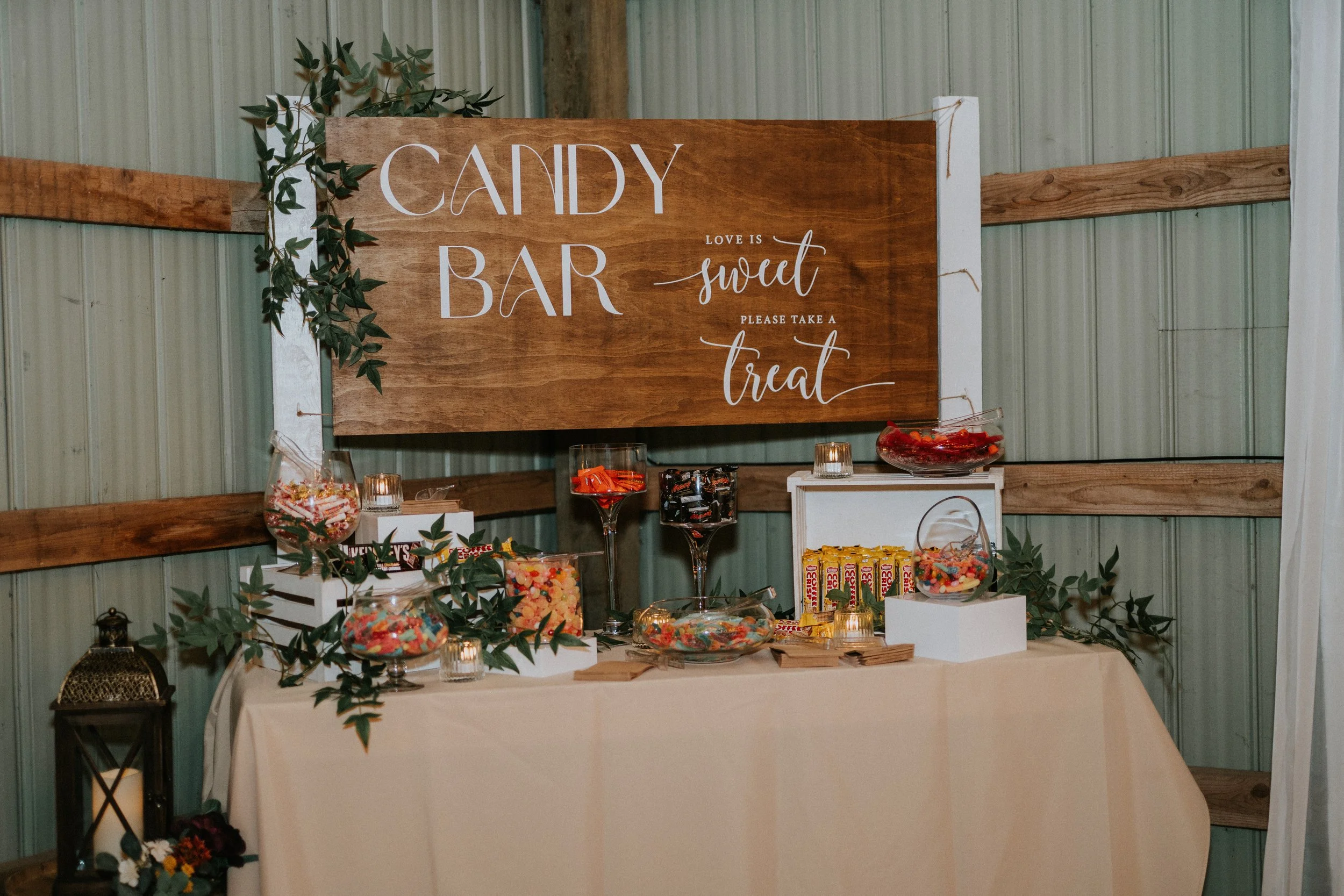 Candy bar setup with a wooden sign reading 'Candy Bar' and 'Love is sweet, please take a treat,' surrounded by containers of colorful candies and decorative greenery.