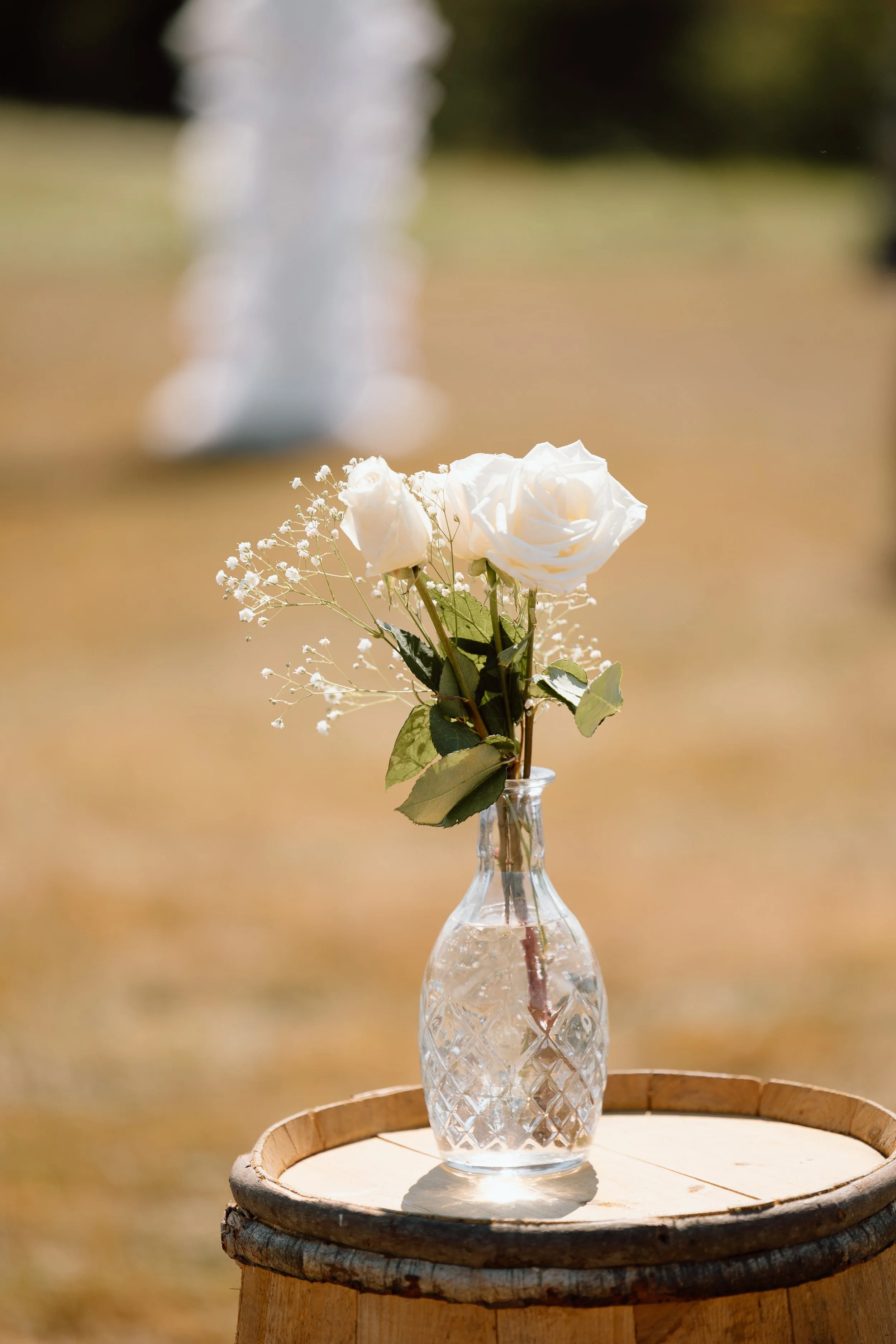 A small glass vase with white roses and baby's breath, placed on a wooden table outdoors during the daytime.