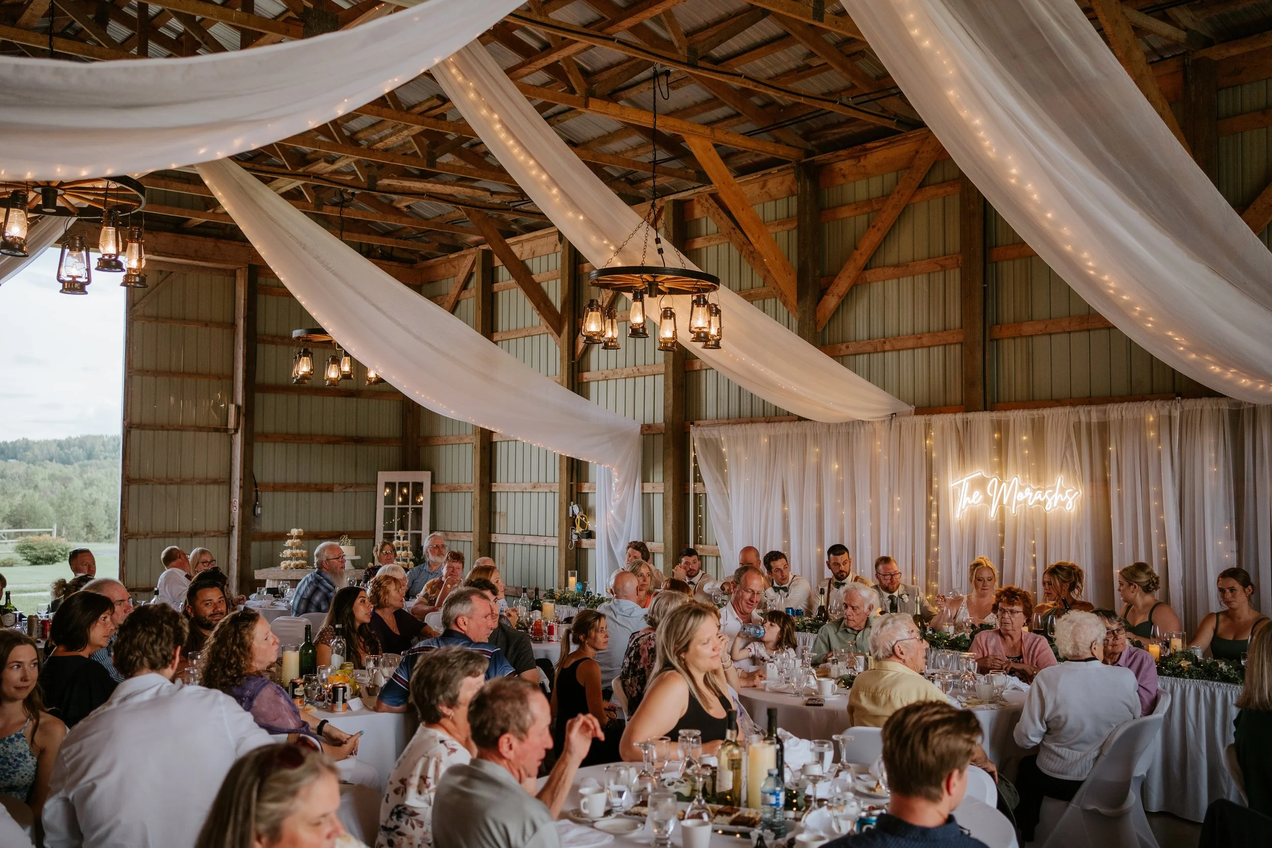 Wedding reception held inside a rustic barn with draped fabric and string lights, featuring guests seated at long tables enjoying food and drinks.