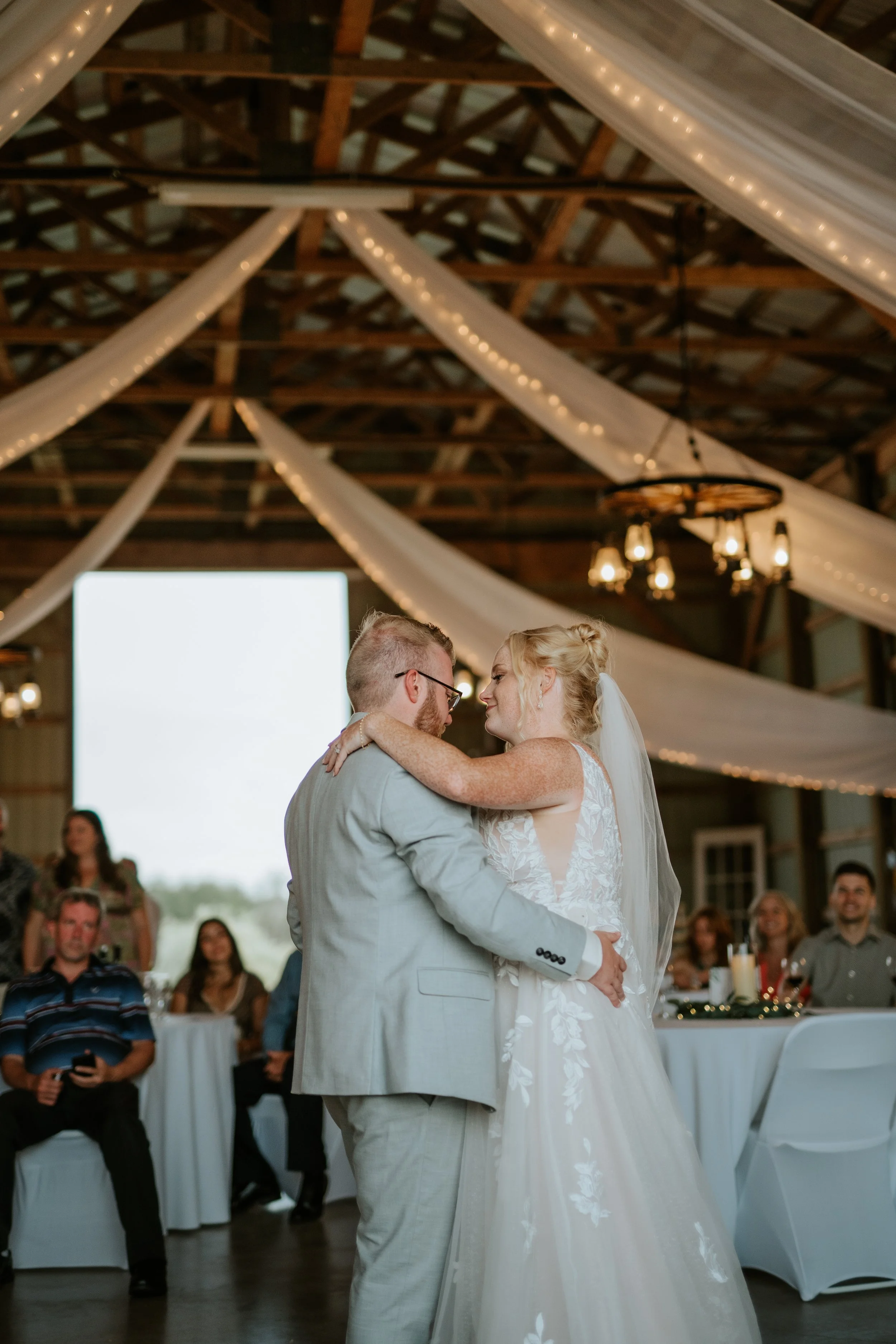 A bride and groom share a dance during their wedding reception in a rustic barn with draped lights and a large window, with guests sitting and watching in the background.