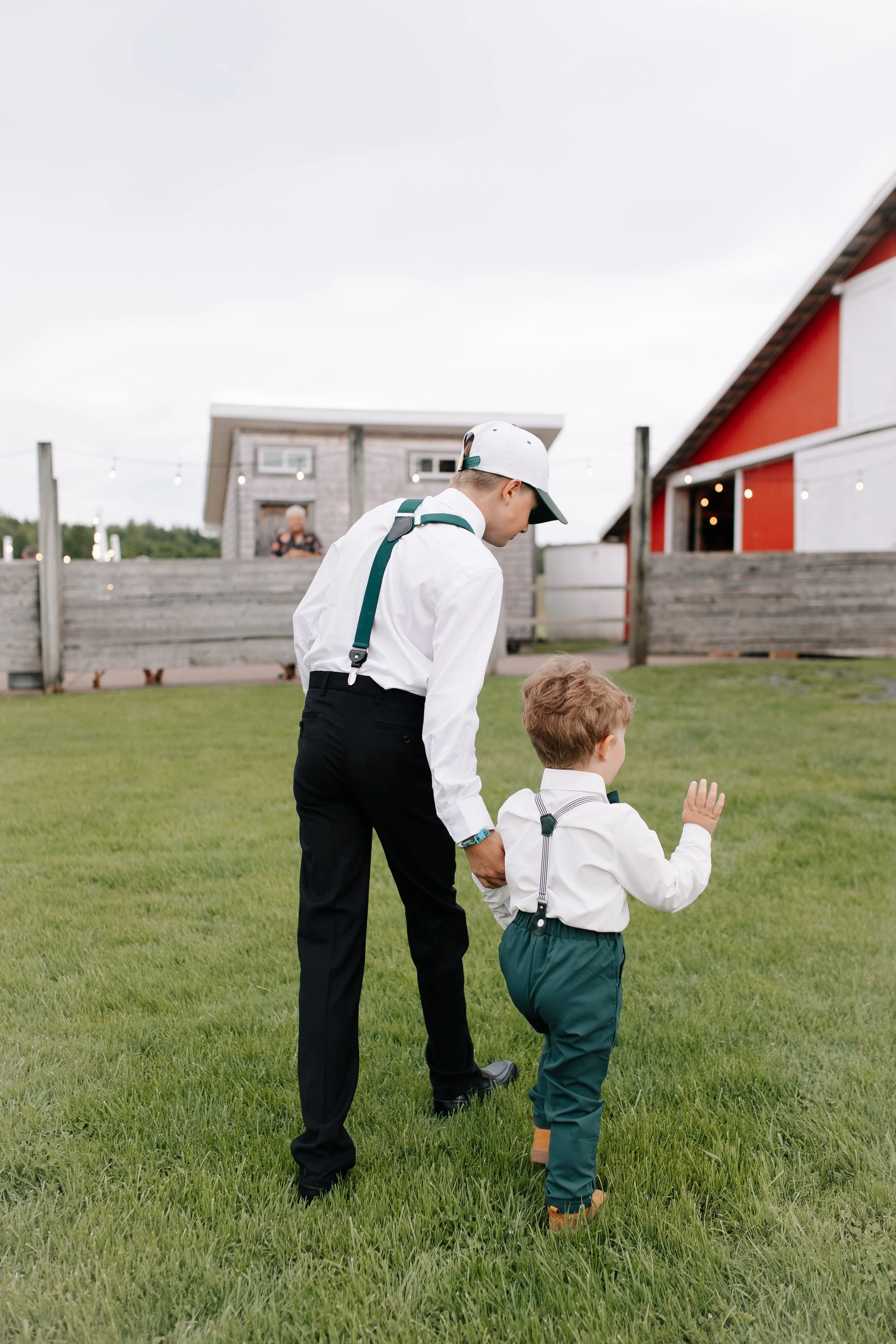 A young man in formal attire holding hands with a young boy, possibly his son, while walking on a grassy field outside. The young man is wearing a white shirt, black pants, and a white cap, and the boy is dressed similarly in a white shirt, darker pa