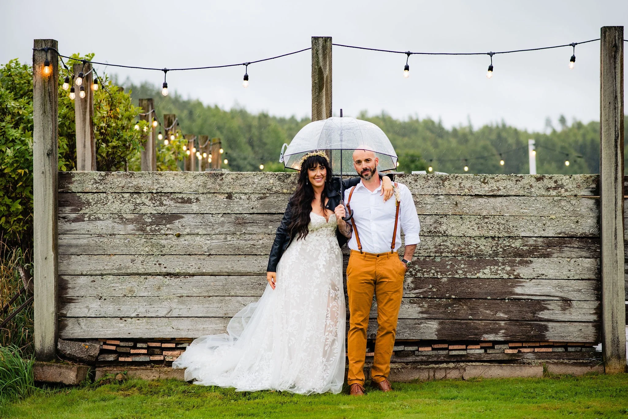 A couple standing together in the rain under a transparent umbrella, with the bride in a white wedding dress and the groom in a white shirt, suspenders, and tan pants, smiling in front of a weathered wooden fence with string lights overhead.