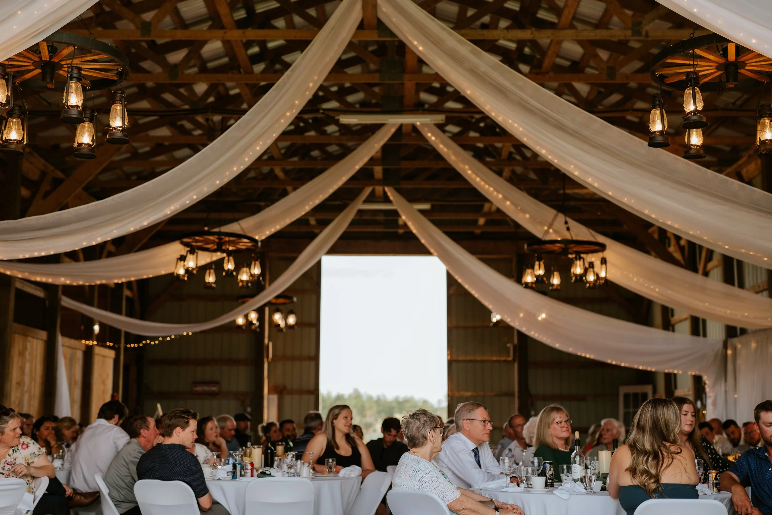 People attending a wedding reception inside a barn with white drapes and chandeliers.