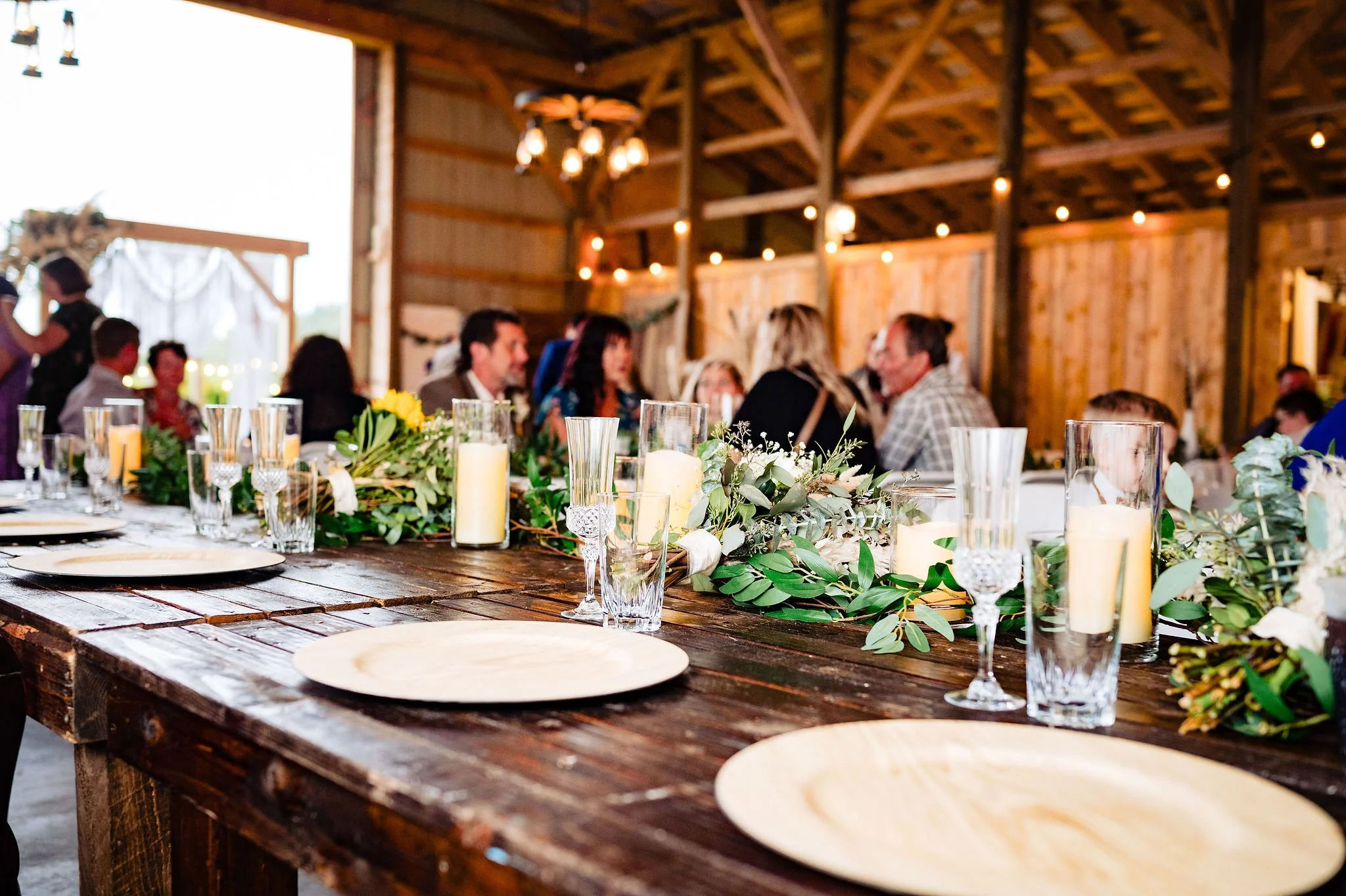 A rustic wedding reception table decorated with greenery, candles, and glassware inside a barn
