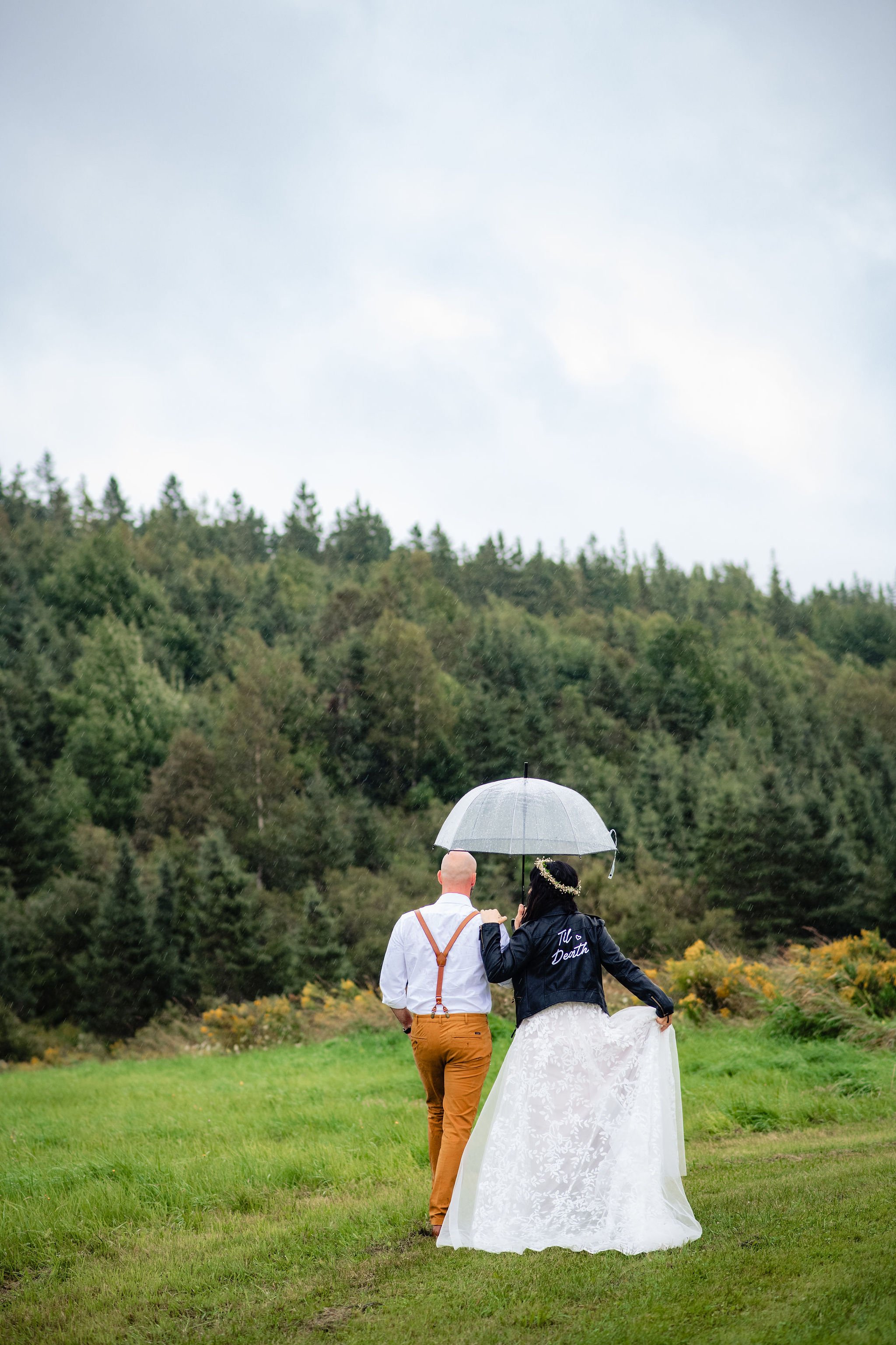A couple walking in a grassy field with trees, sharing an umbrella on a rainy day at a wedding or special event.